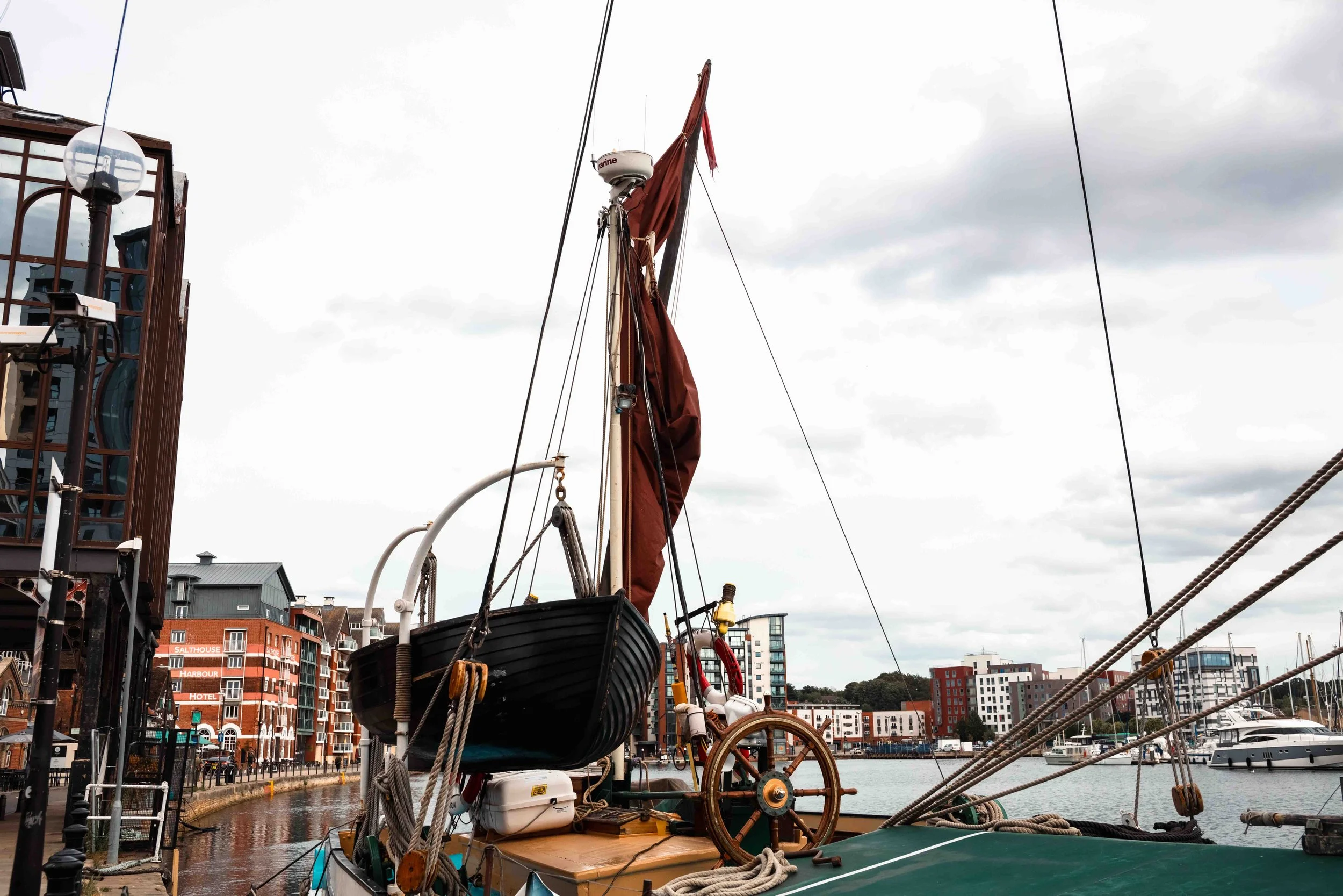 A boat docked at a harbor with modern buildings, including a hotel, visible in the background. The boat has a tall mast with a furled sail and is surrounded by other boats on the water, under an overcast sky.