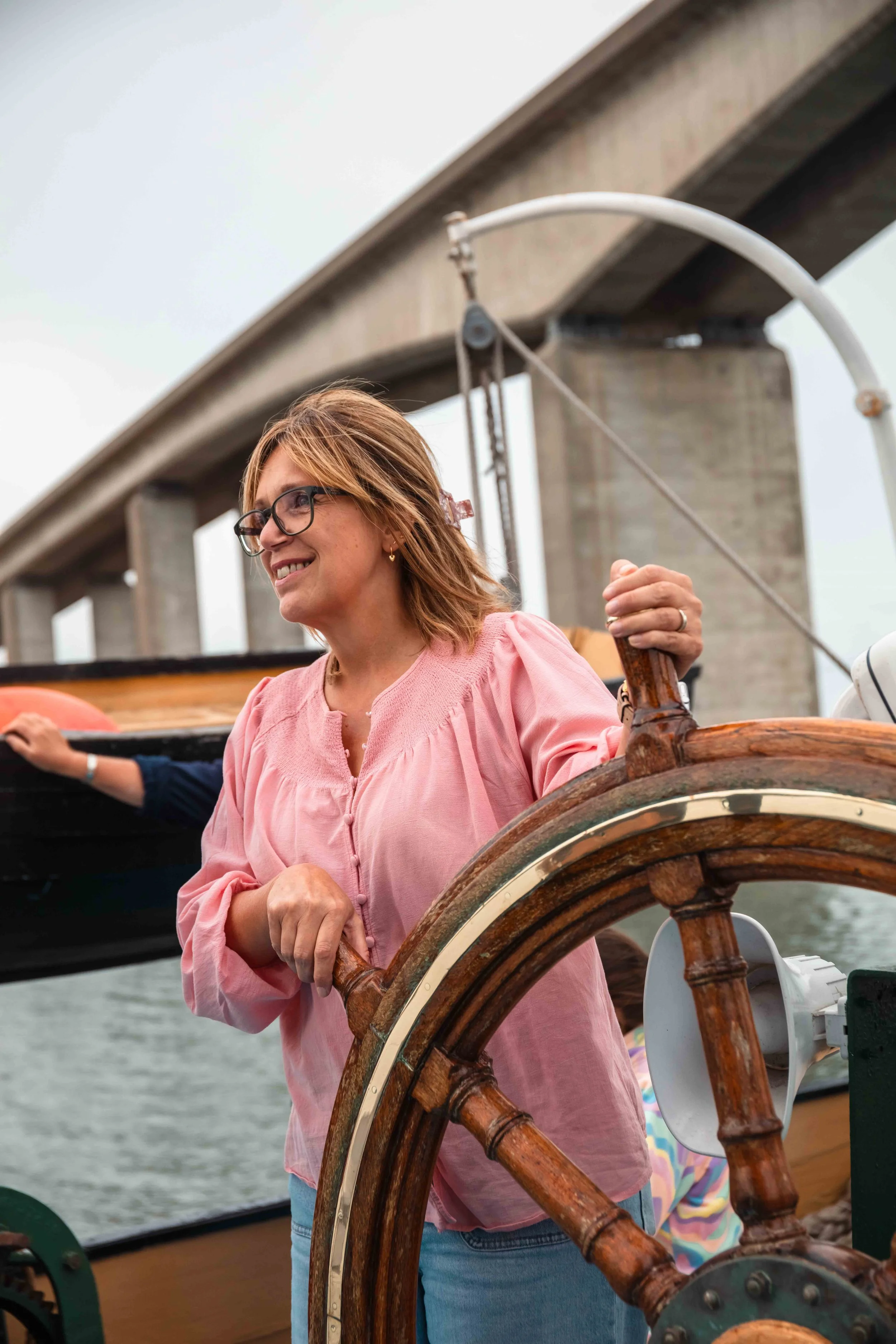 Woman smiling and steering a boat, with a bridge in the background.