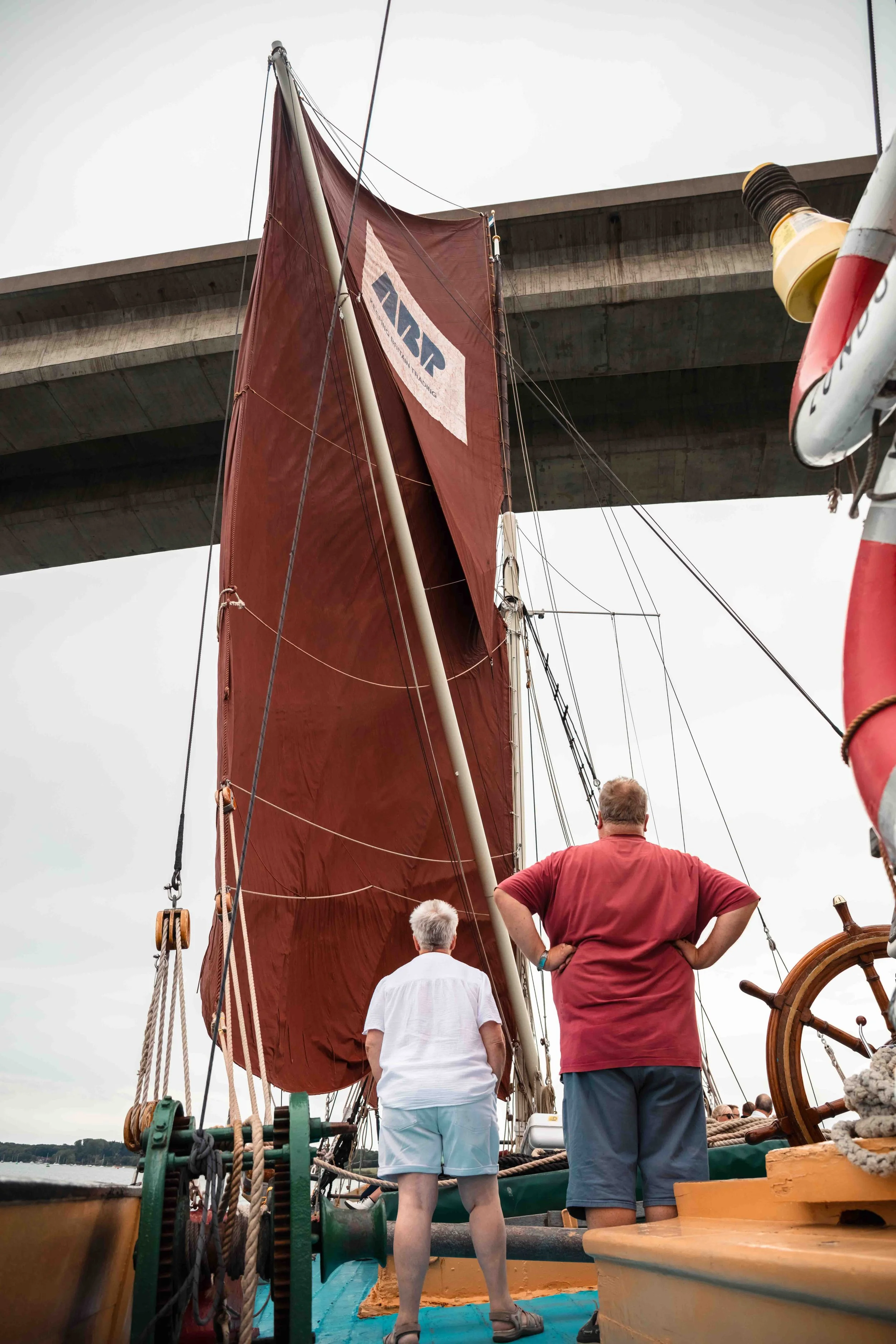 People aboard a sailboat, looking up at its large red sail and a bridge overhead, with an overcast sky.