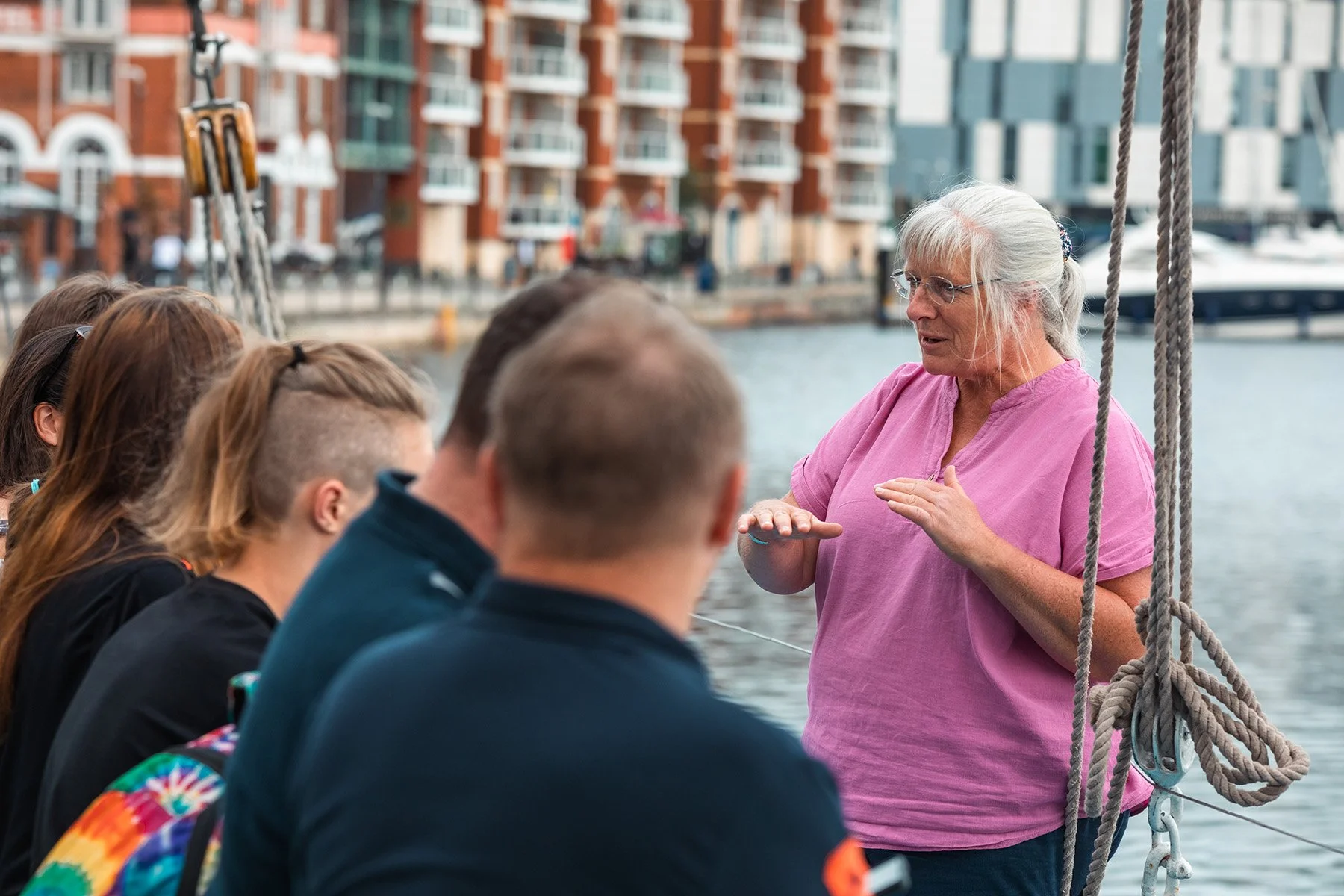 An elderly woman with white hair and glasses giving a talk to a group of young people on a boat by the water in an urban area.