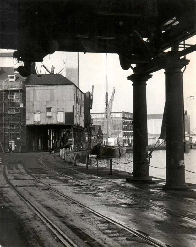 Black and white photo of an old harbor scene with ship, warehouses, and railway tracks under a bridge with large supporting columns.