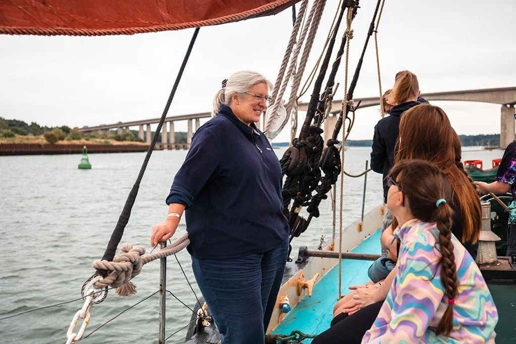 A group of people on a sailboat, with a woman standing and talking to others, on a body of water with a bridge in the background.