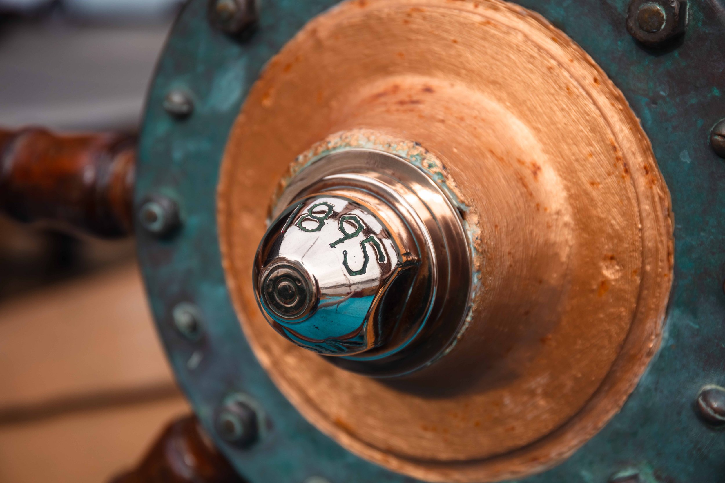 Close-up of a shiny 1896 vintage mechanical timer or gauge attached to a copper-colored cylindrical part, mounted on a weathered teal metal surface with rusted bolts.