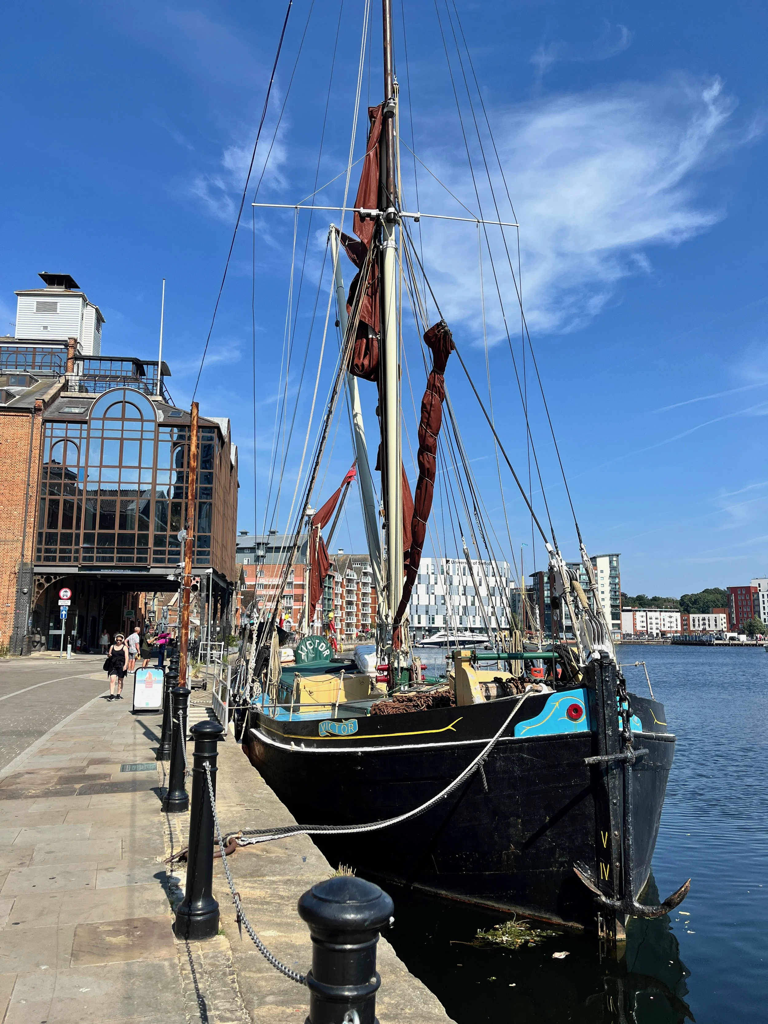 A black sailboat named Victor docked along a city waterfront, with buildings and people walking on the promenade, under a partly cloudy blue sky.