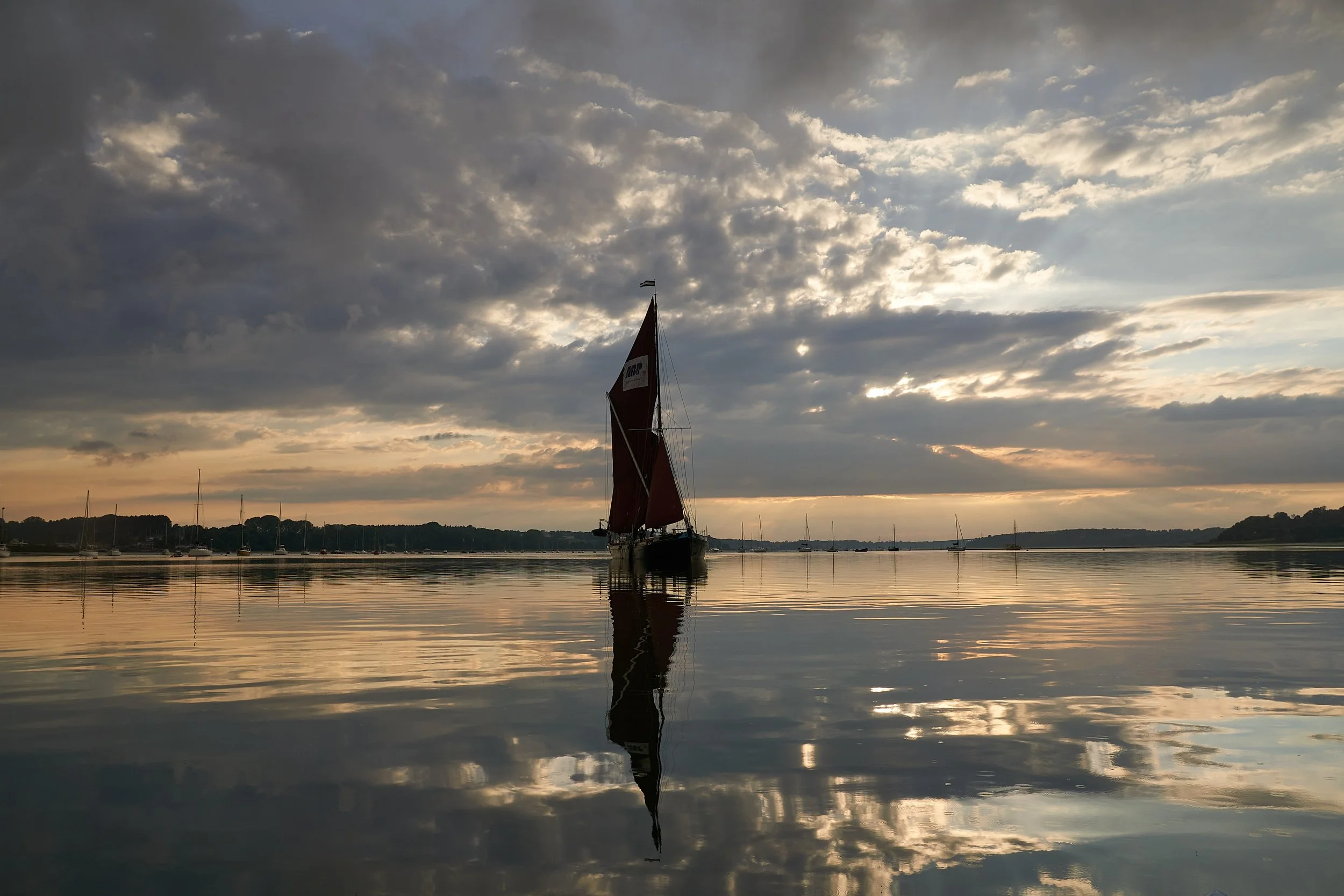 A sailboat with dark sails sailing on calm water during sunset, with a cloudy sky and distant shoreline.