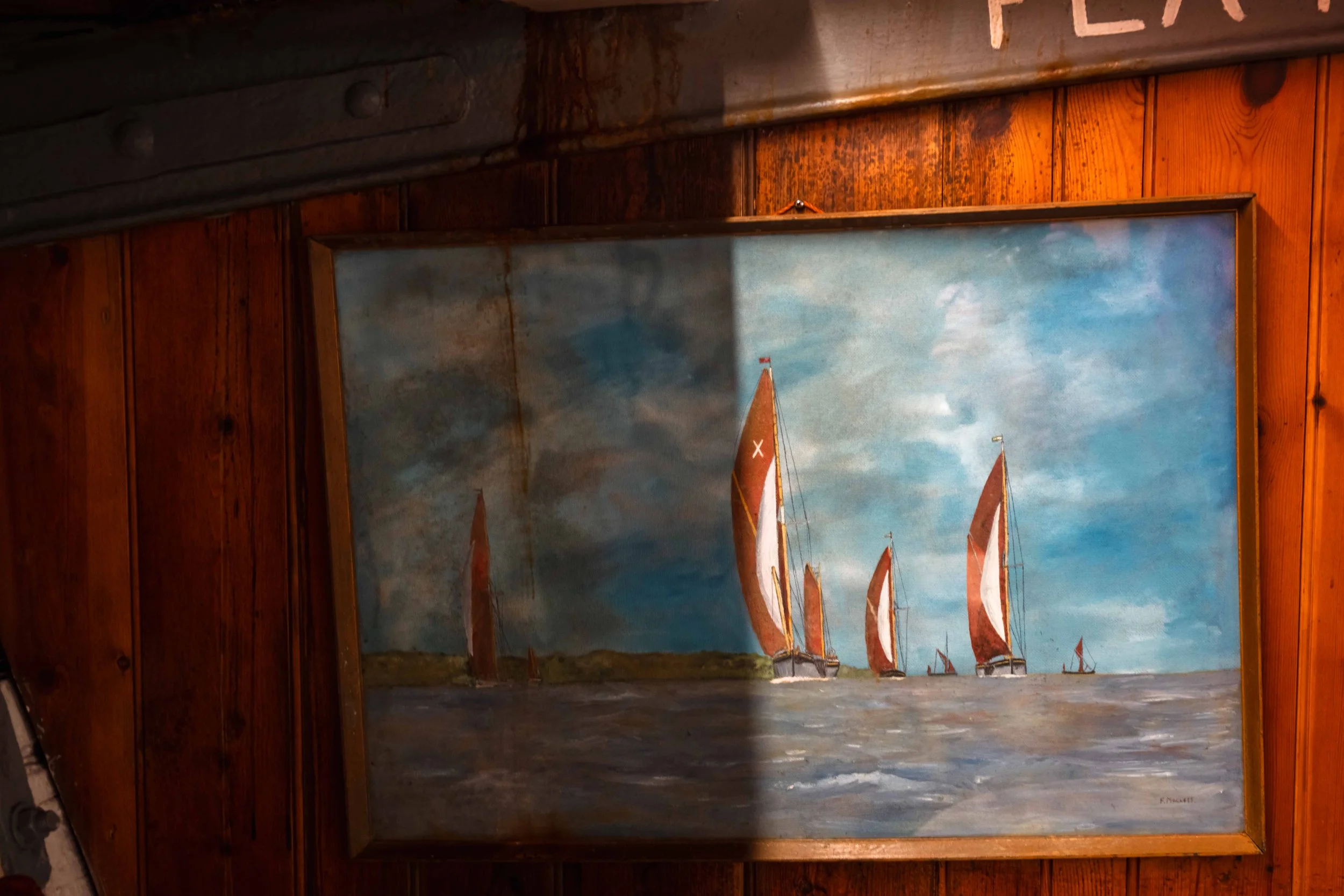A painting of sailboats with red and white sails on the water under a partly cloudy sky, framed and hung on a wooden wall.
