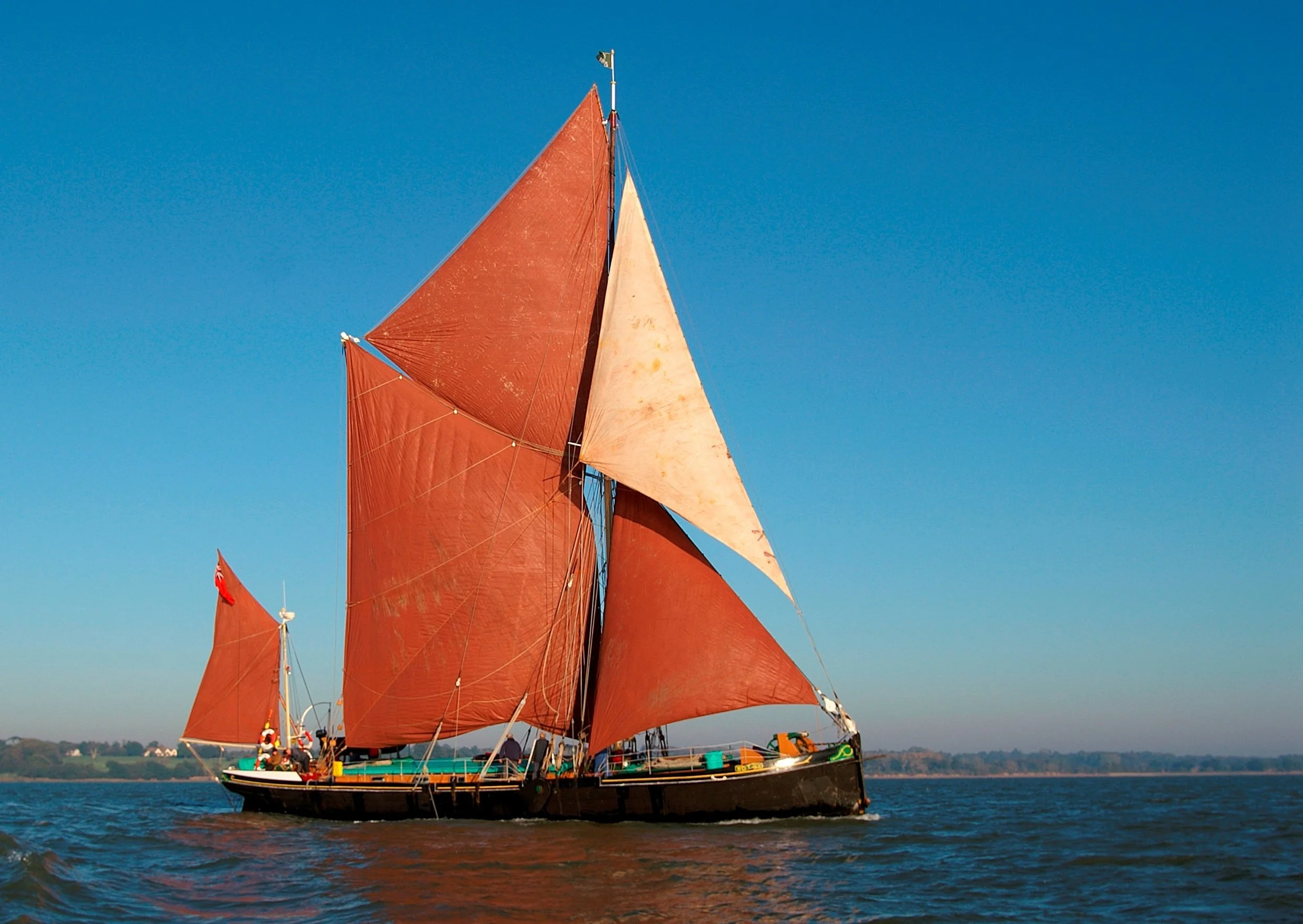 A traditional sailing ship with bright red sails on a body of water, with a clear blue sky and a distant shoreline.