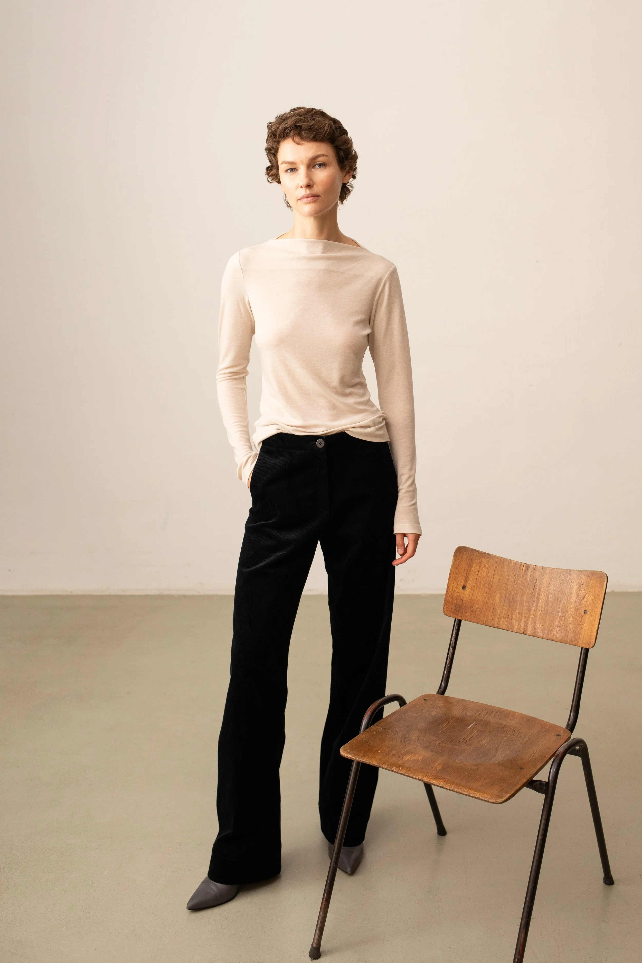 Eine junge Frau mit kurzen, lockigen Haaren steht in einem minimalistischen Raum vor einer weißen Wand. Sie trägt eine beige lange Ärmel Bluse, schwarze Hosen und graue Schuhe. Neben ihr steht ein einfaches Holzstuhl mit Metallgestell.