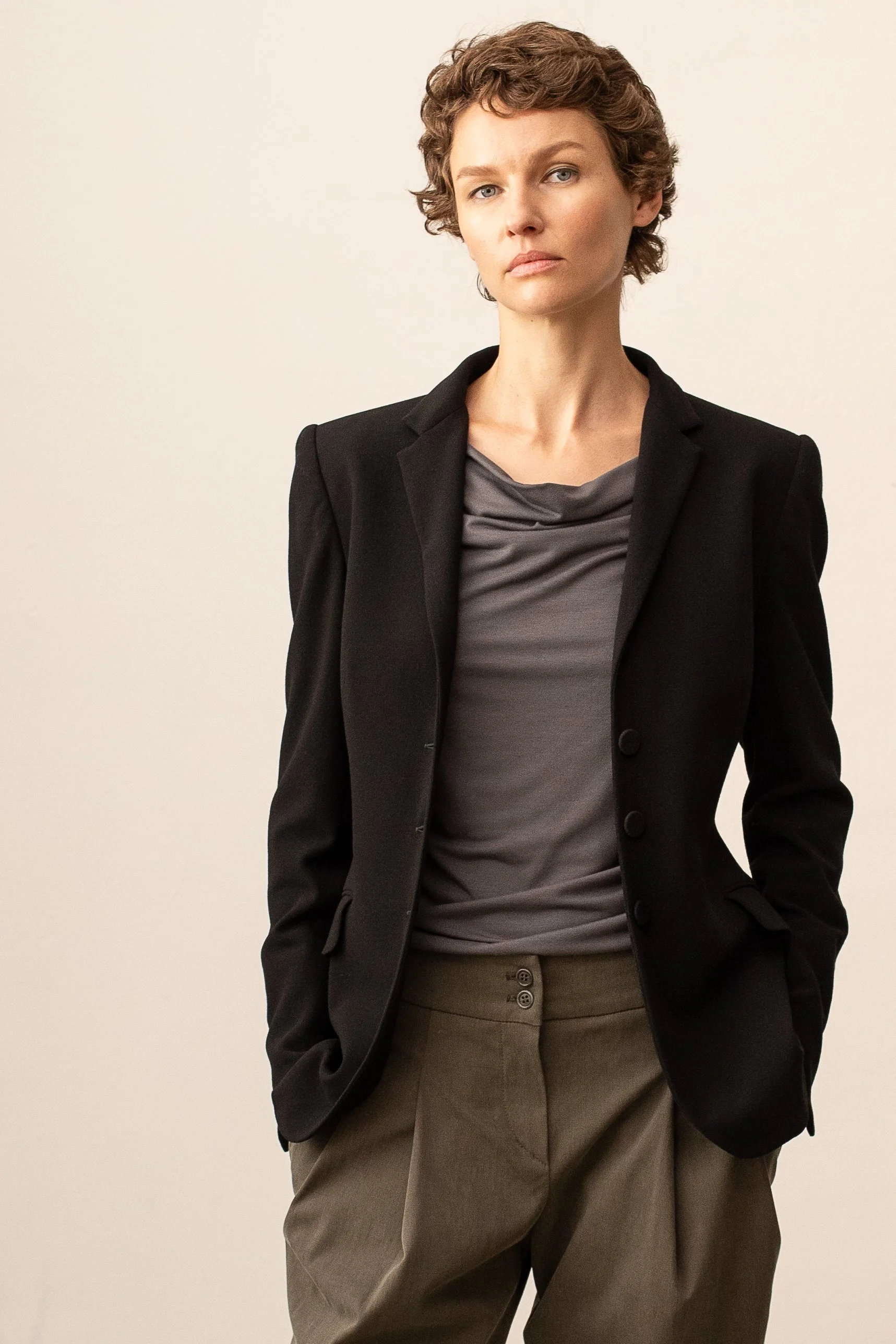 Eine Frau mit kurzen braunen Haaren trägt eine schwarze Blazerjacke, ein graues Oberteil und braune Hosen, steht vor einem cremefarbenen Hintergrund.