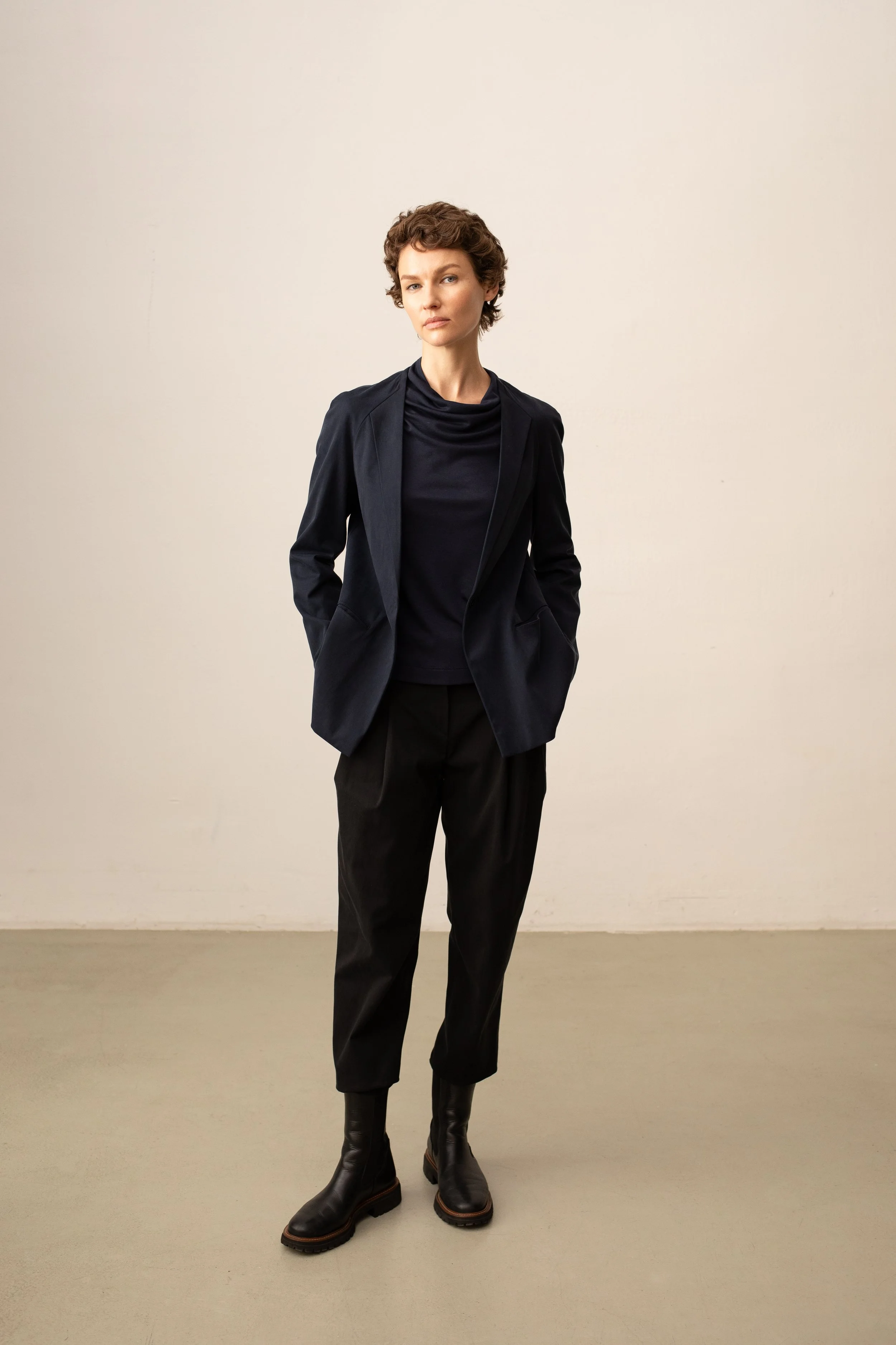 Eine Frau mit kurzen braunen Haaren trägt eine schwarze Bluse, eine schwarze Jacke, schwarze Hosen und schwarze Stiefel, steht vor einer weißen Wand.