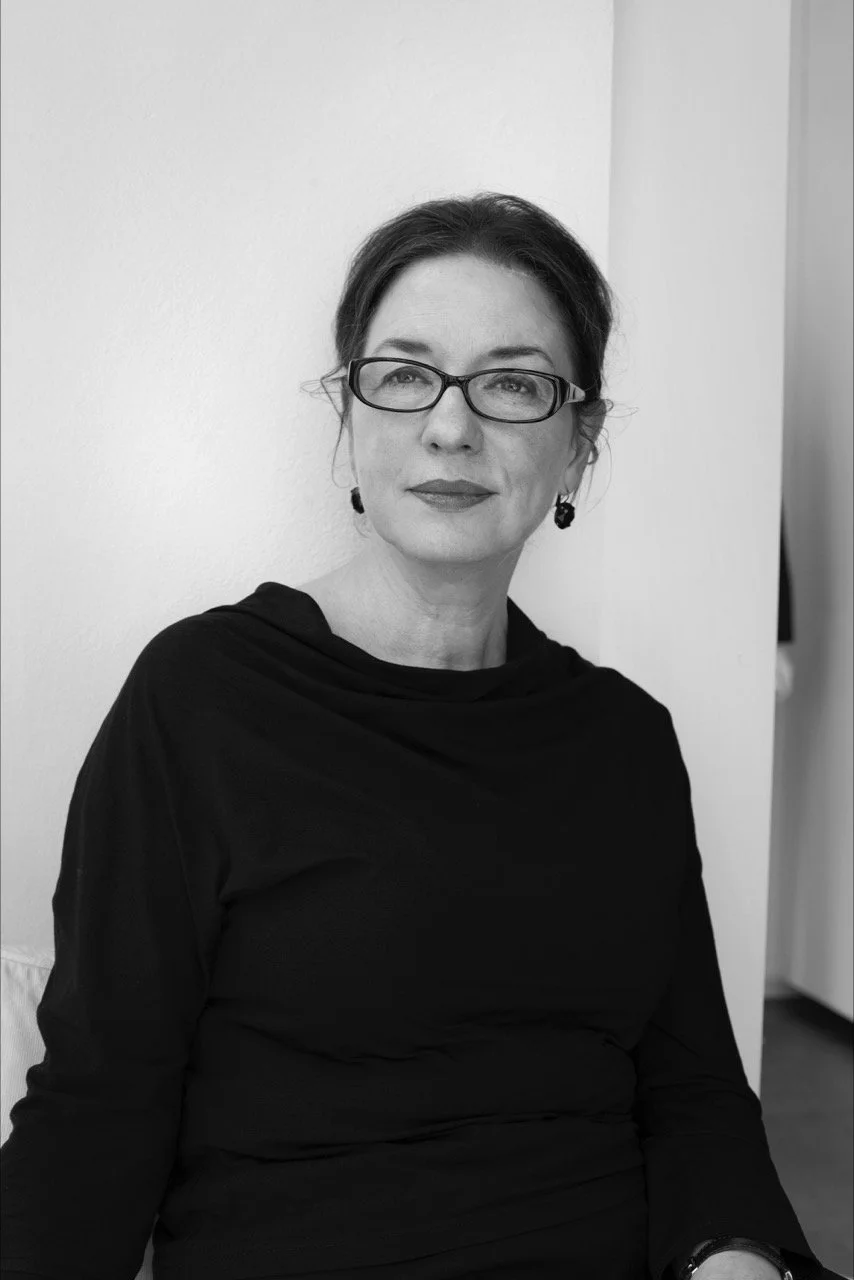 Schwarz-weiß Foto einer Frau mit dunklem Haar, schwarzen Brillen, roten Lippen, Ohrringen und schwarzem Kleid, sitzend vor weißer Wand.