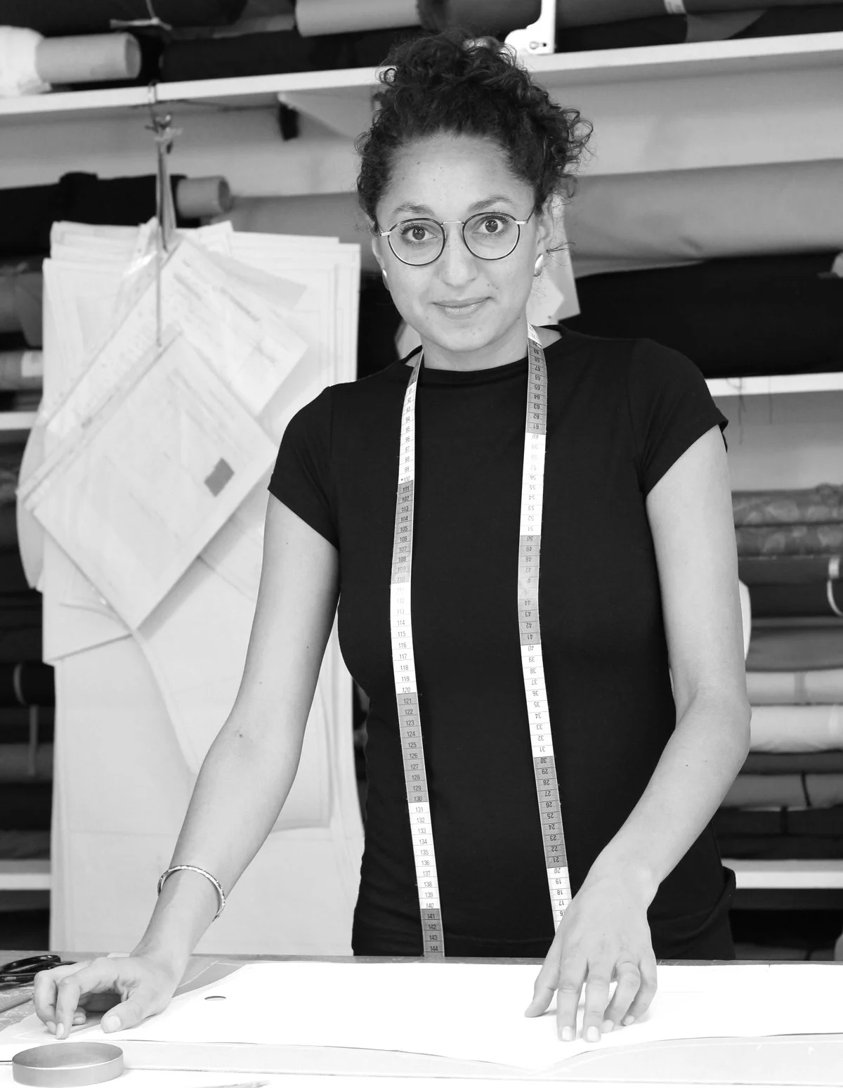 Eine Frau in einem Schneideratelier steht vor einem Tisch, trägt eine Messlatte um den Hals und arbeitet an einem Stoffstück.