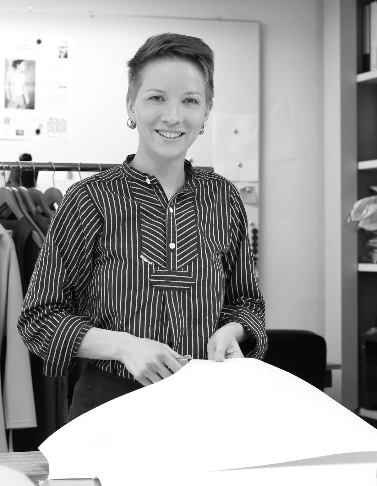 Schwarzes und weißes Foto einer Frau mit kurzen Haaren, die in einem Büro steht, mit Hängekleidung im Hintergrund, und eine Aufgabe mit Papier erledigt.
