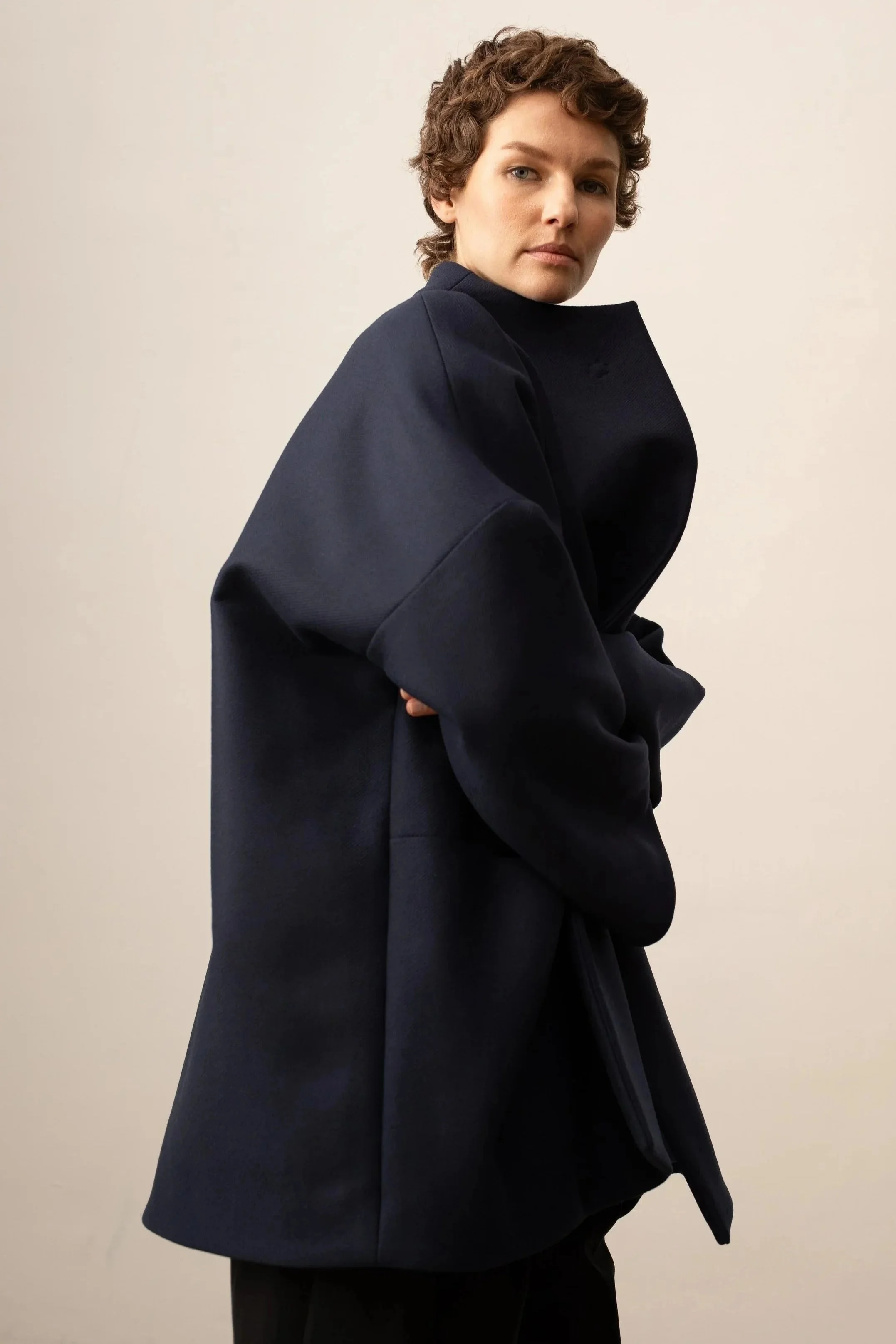 Porträt einer jungen Frau mit kurzen, lockigen braunen Haaren, in einem schwarzen Pullover vor einem hellen Hintergrund.