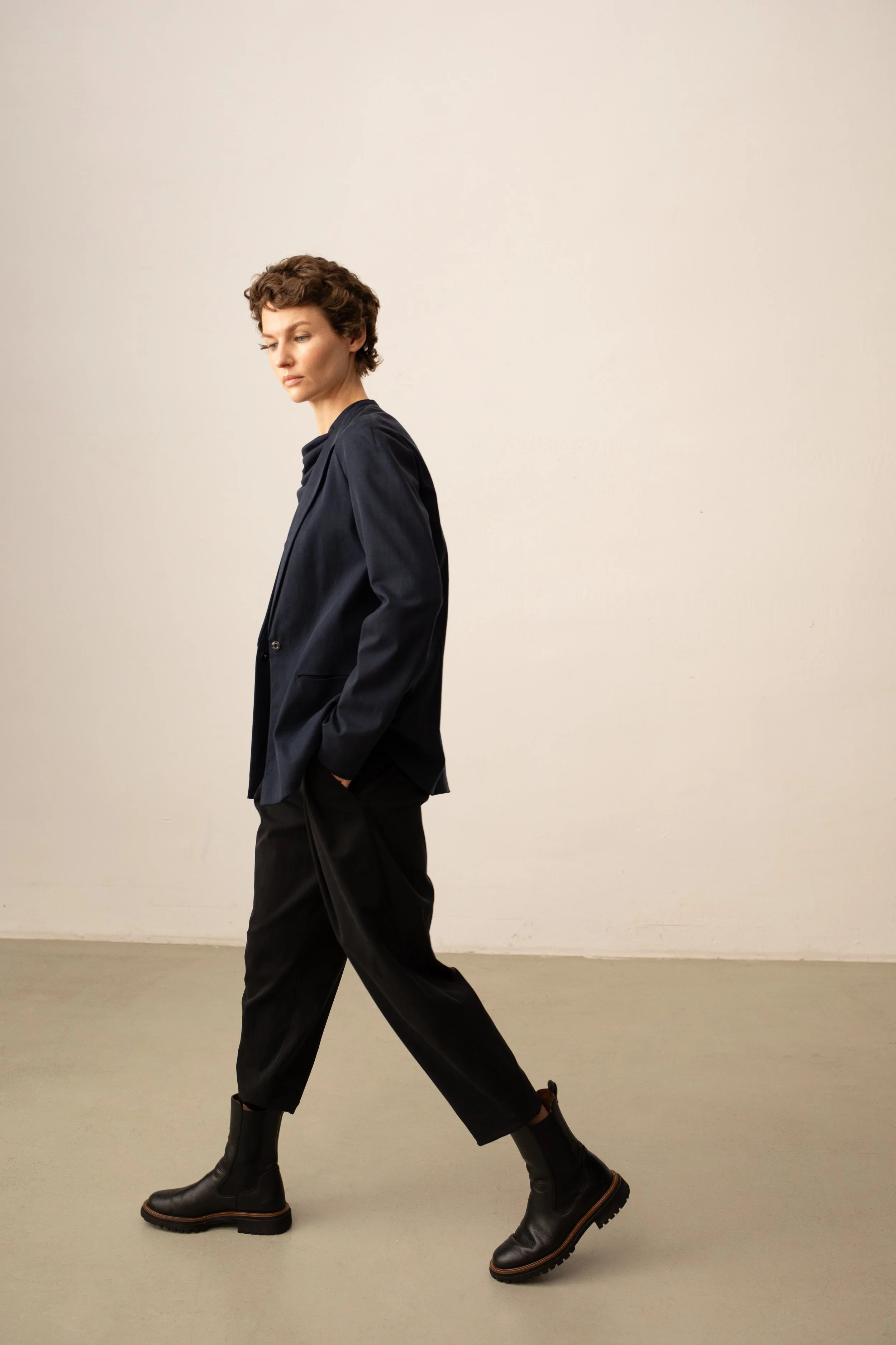 Eine Frau mit kurzen, lockigen braunen Haaren trägt eine schwarze Jacke, schwarze Hose und schwarze Stiefel, während sie in einem minimalistischen, hellen Raum langsam geht.