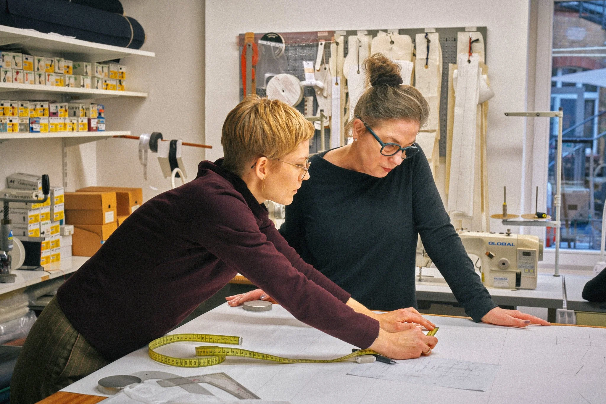 Zwei Frauen arbeiten in einer Design- oder Schneiderei-werkstatt und messen Stoffe mit einem Maßband auf einem Tisch