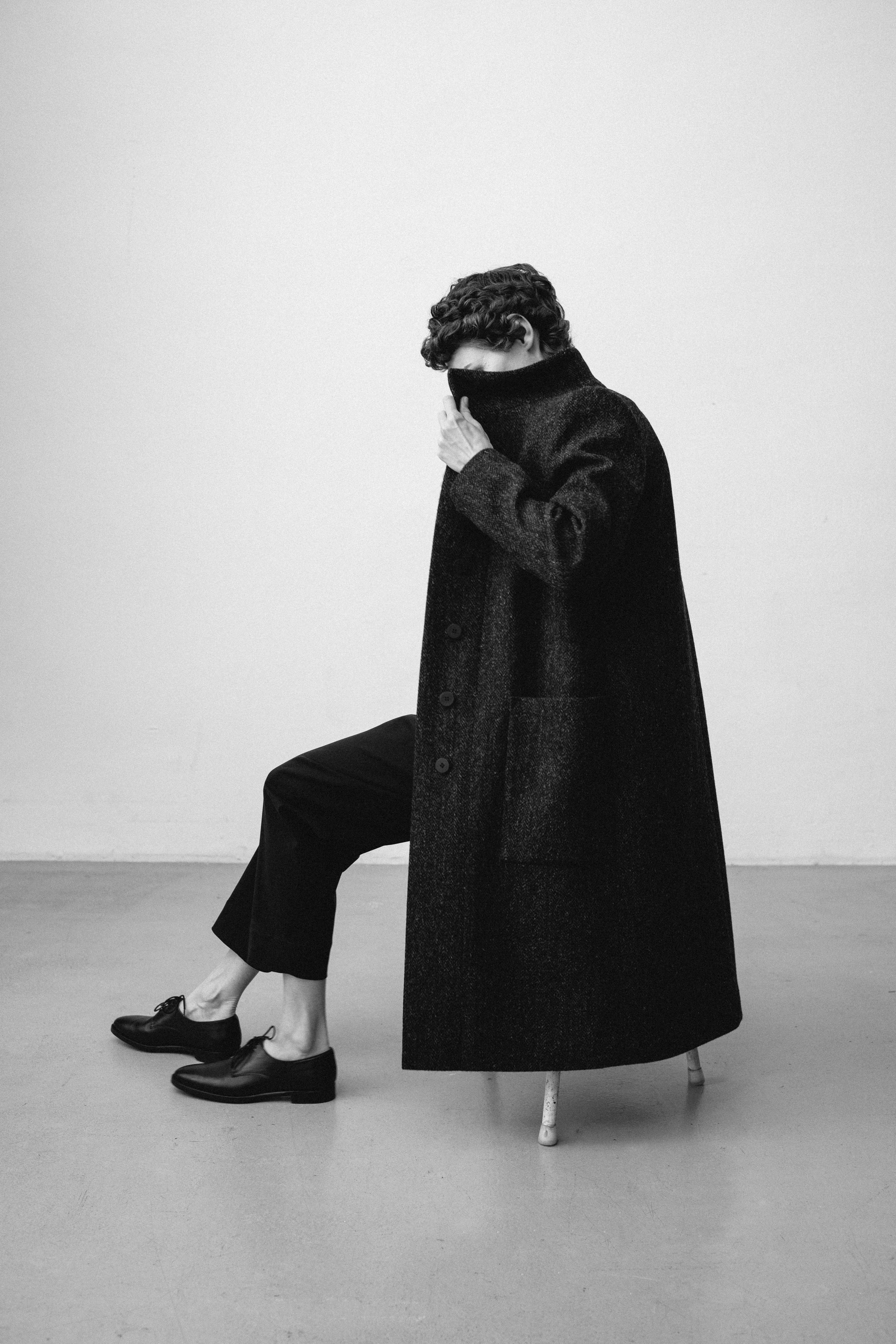 Schwarz-weiß-Foto einer Person mit Locken, die in einen schwarzen Pullover mit hohem Kragen eingetaucht ist, der das Gesicht bedeckt. Die Person sitzt auf einem Stuhl mit gebrochenem Bein, trägt schwarze Schuhe und schwarze Hosen. Der Hintergrund ist schlicht und hell.