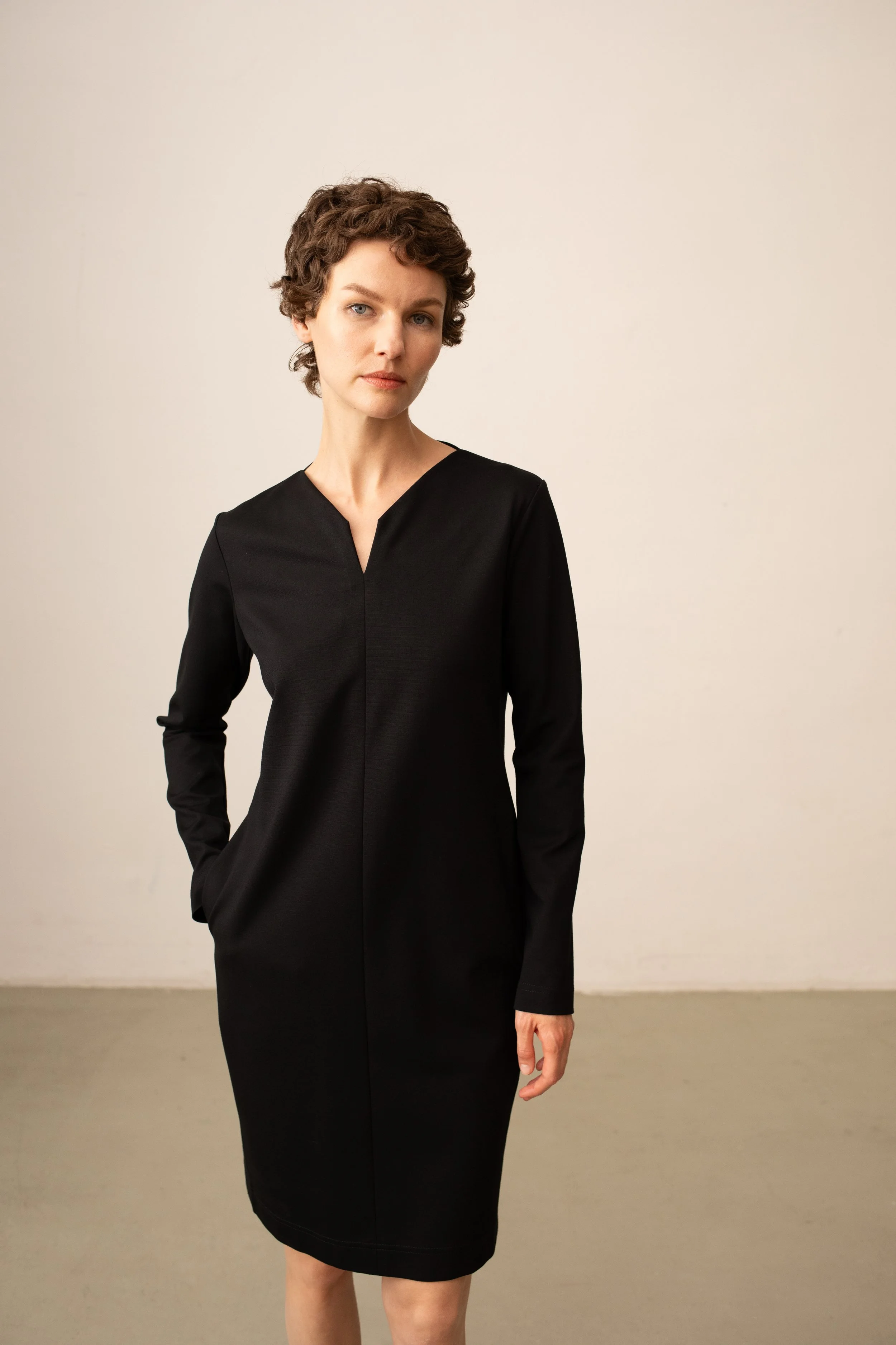 Porträt einer Frau mit kurzen, lockigen braunen Haaren, in einem schwarzen Kleid, steht vor neutralem Hintergrund.