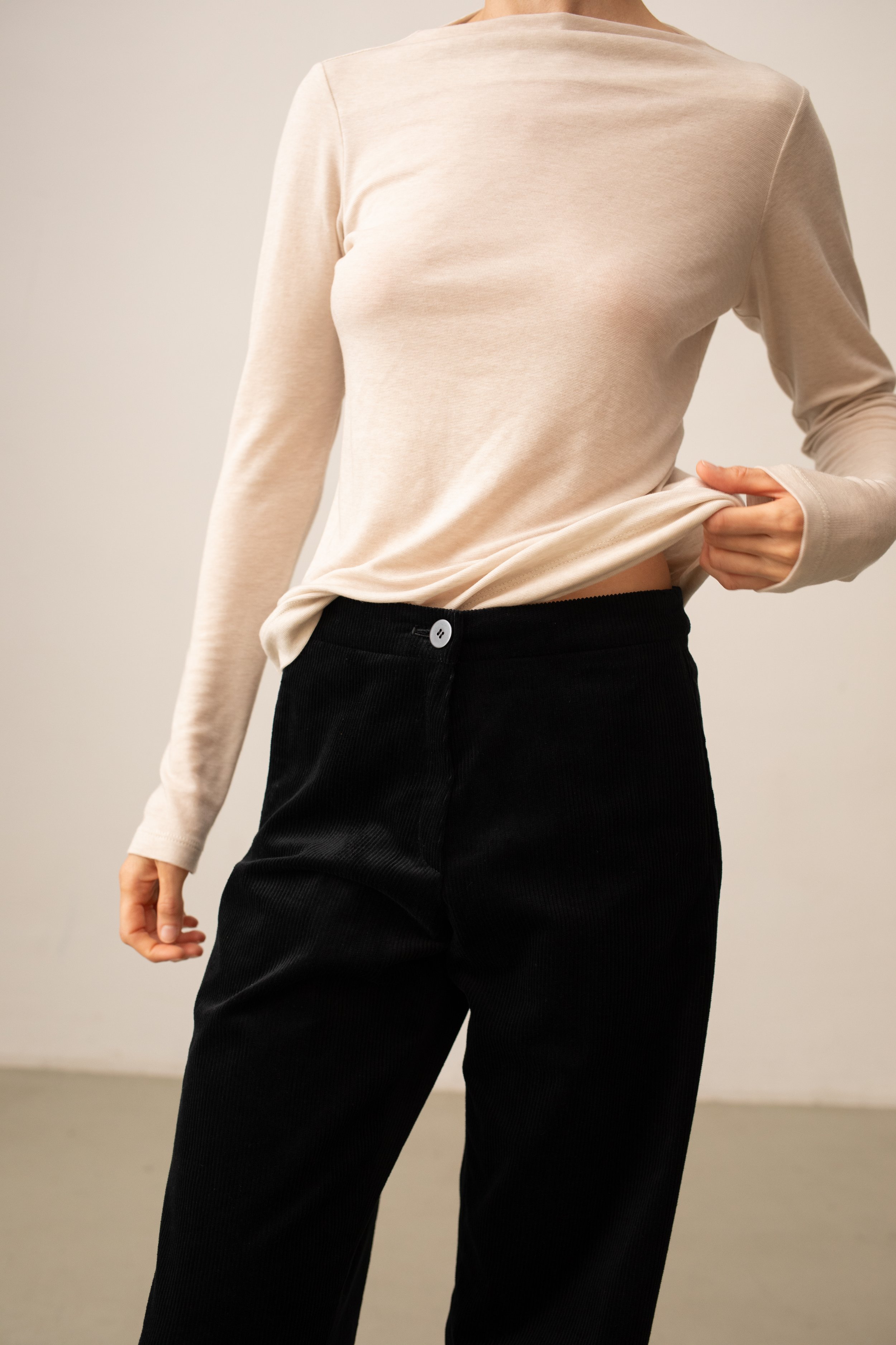 Nahaufnahme eines Menschen, der beige Pullover trägt, der leicht an der Taille hochgezogen wird, und schwarze Hosen. Der Hintergrund ist neutral.