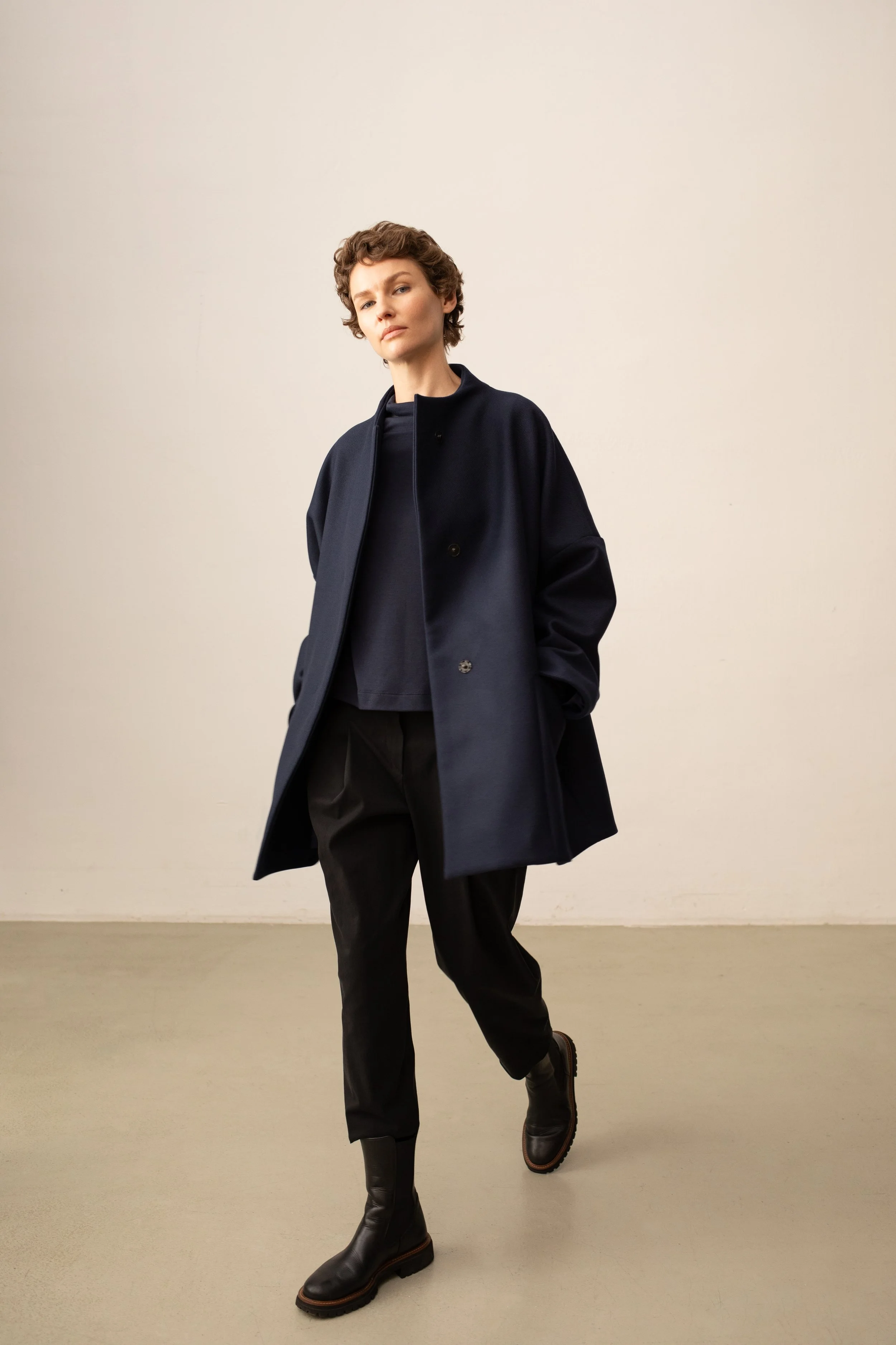 Eine Frau mit kurzen, lockigen braunen Haaren trägt eine dunkle Jacke, eine Bluse und schwarze Hose. Sie steht vor einer neutralen Wand, schaut ernst und selbstbewusst in die Kamera, mit einer Hand in der Tasche und einem Bein leicht vor dem anderen.
