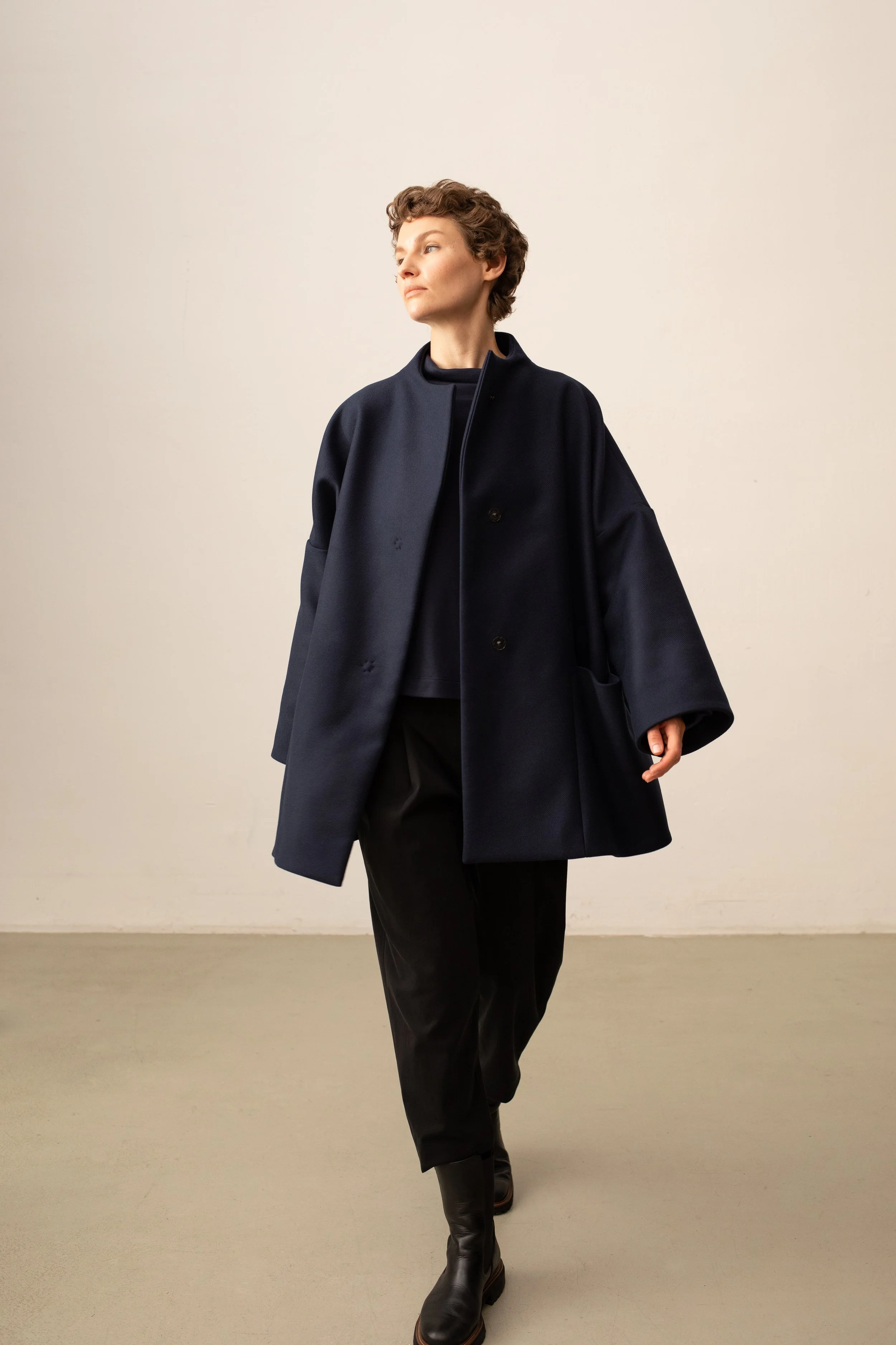 Eine Frau mit kurzes, lockigem braunem Haar trägt einen schwarzen Mantel, schwarze Hose und kniehohe schwarze Stiefel vor einem neutralen, hellen Hintergrund.