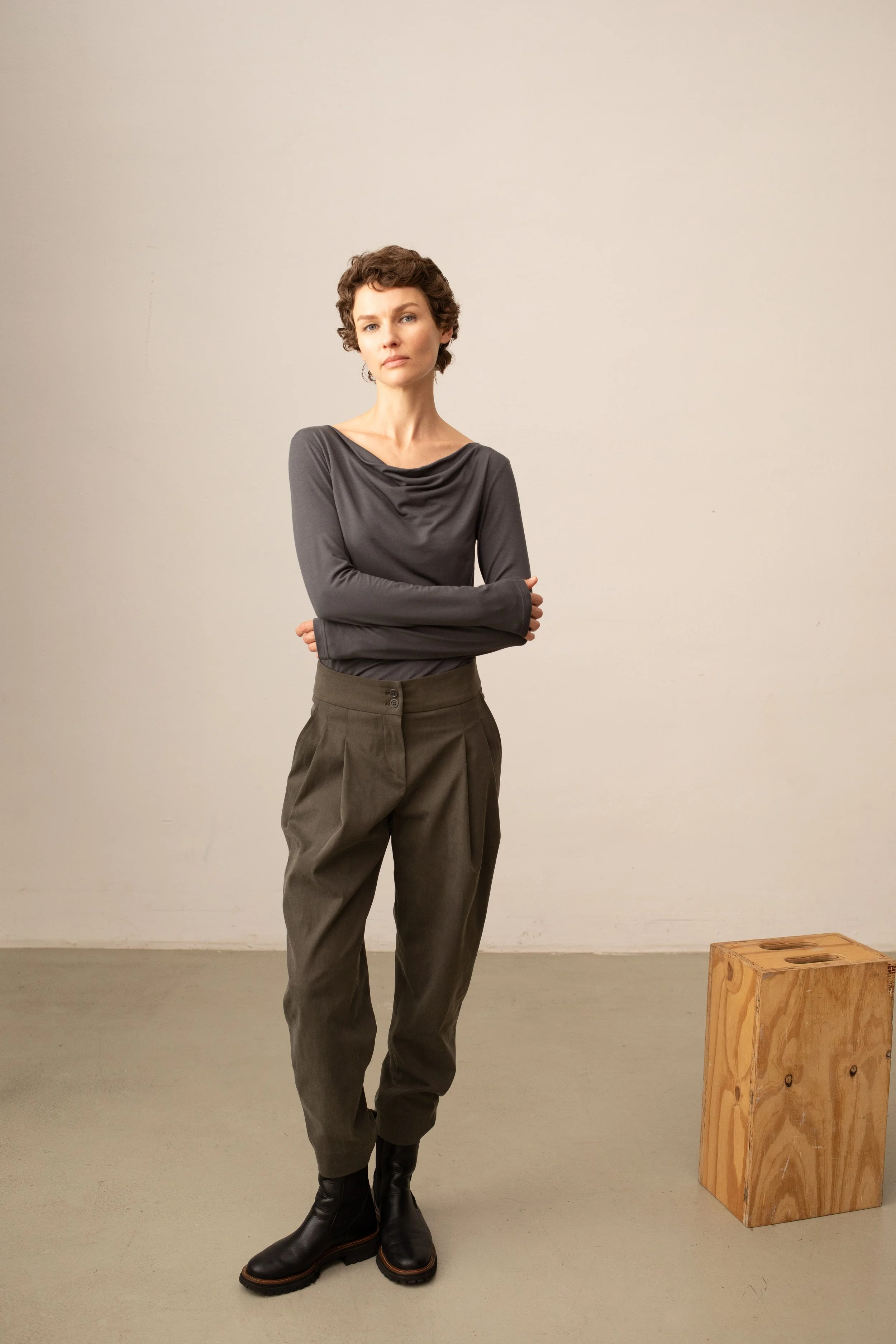 Eine Frau mit kurzen, lockigen braunen Haaren, trägt eine dunkle graue Bluse und braune, weite Hosen, schwarze Stiefel, steht mit verschränkten Armen vor einer schlichten Wand neben einem Holzkasten.
