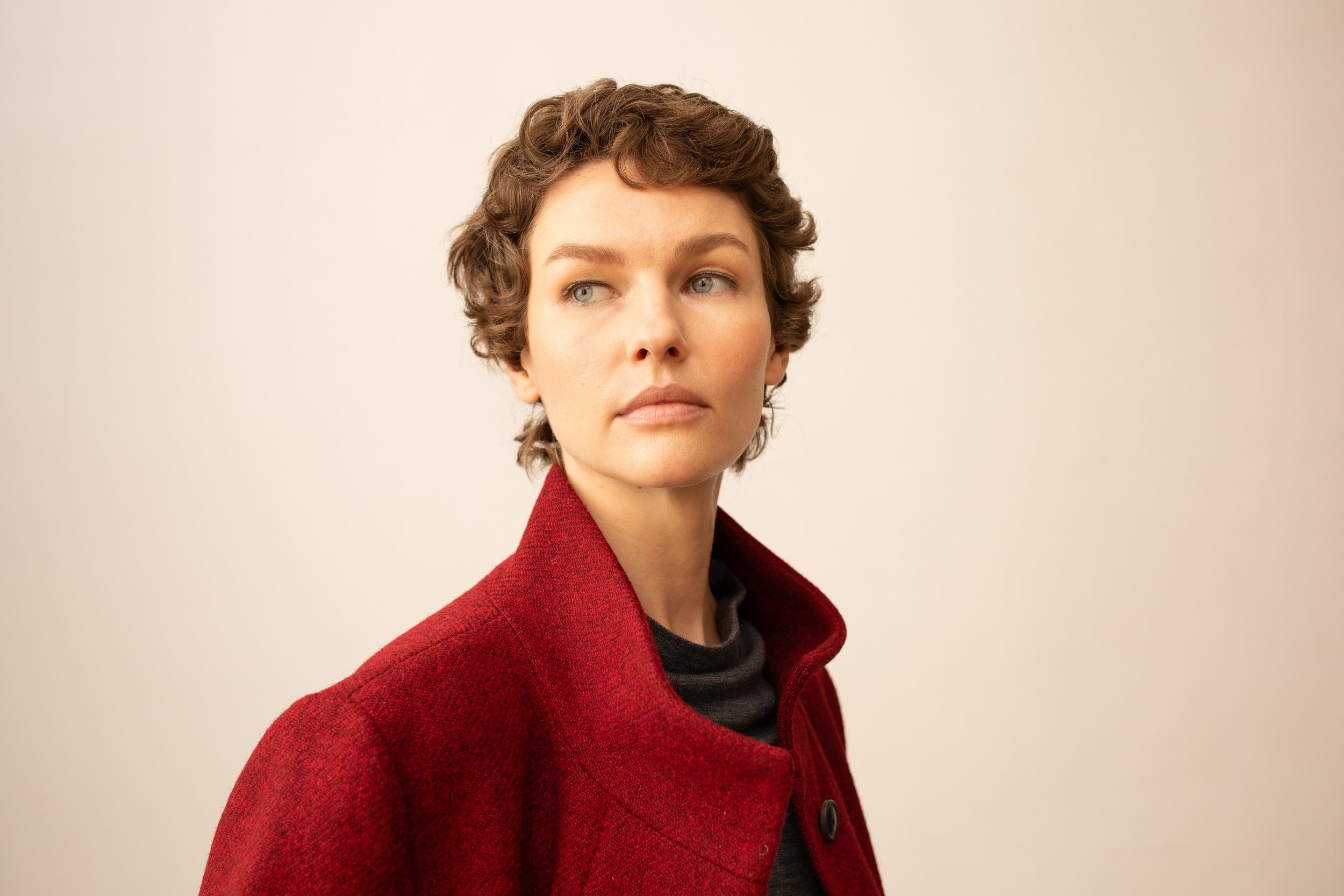 Porträt einer Frau mit kurzen, lockigen braunen Haaren, blauen Augen, trägt einen roten Mantel und eine graue Strickjacke vor einem neutralen Hintergrund.