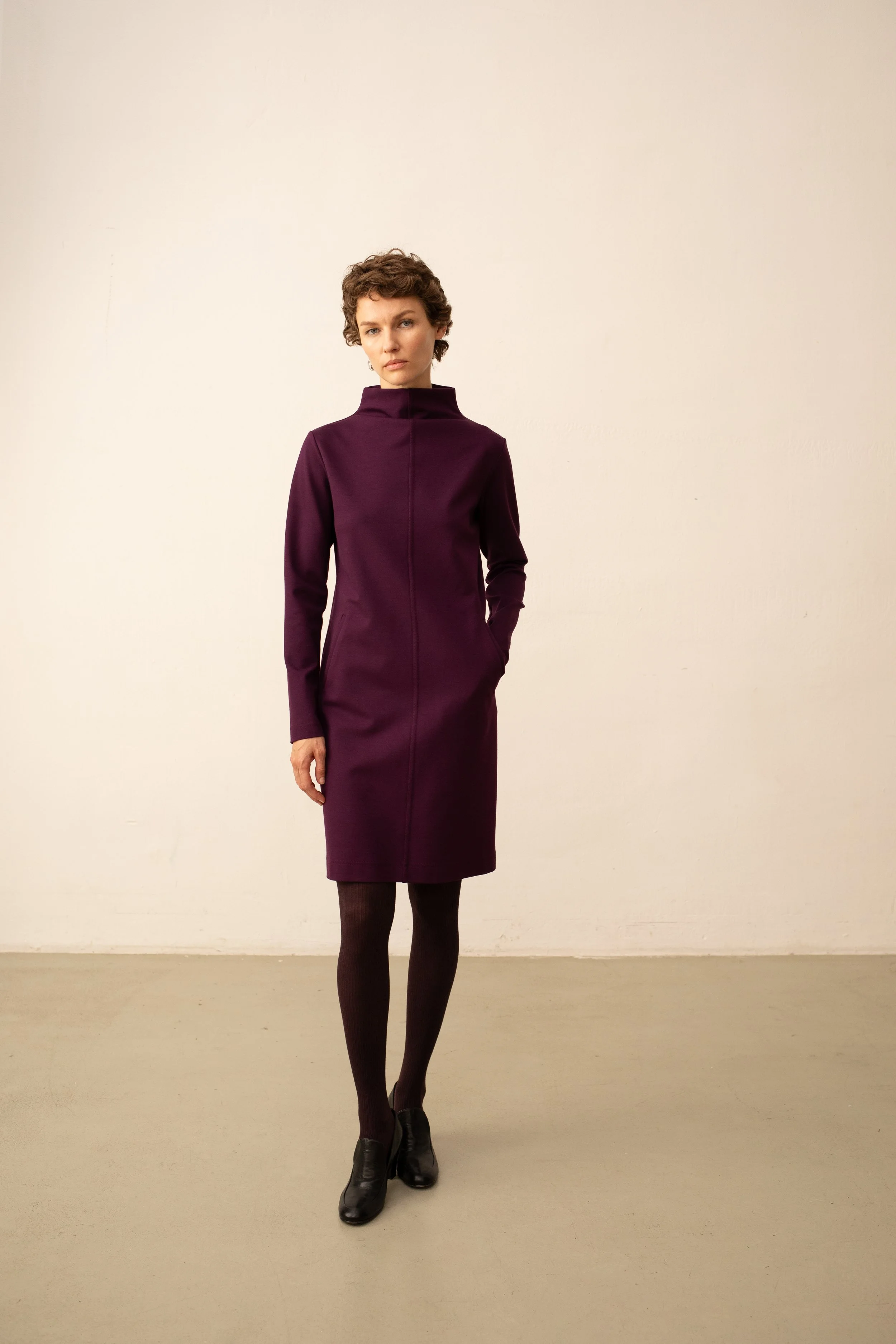 Eine Frau steht vor einer weißen Wand, trägt ein dunkellila, knielanges Kleid mit hohem Kragen, schwarze Strumpfhosen und schwarze Schuhe.