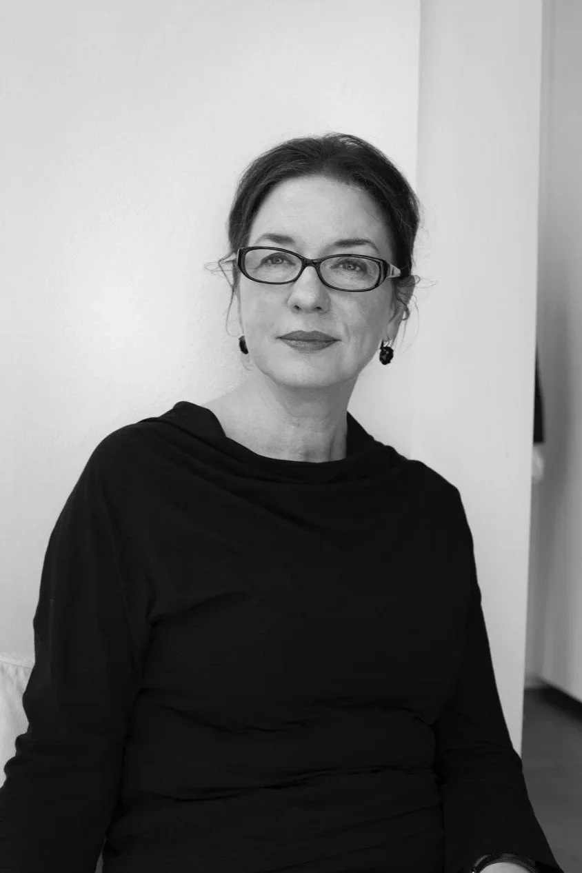 Schwarz-weiß-Foto einer Frau mit dunklen Haaren, Brille und dunklem Kleid, sitzend an einer Wand.