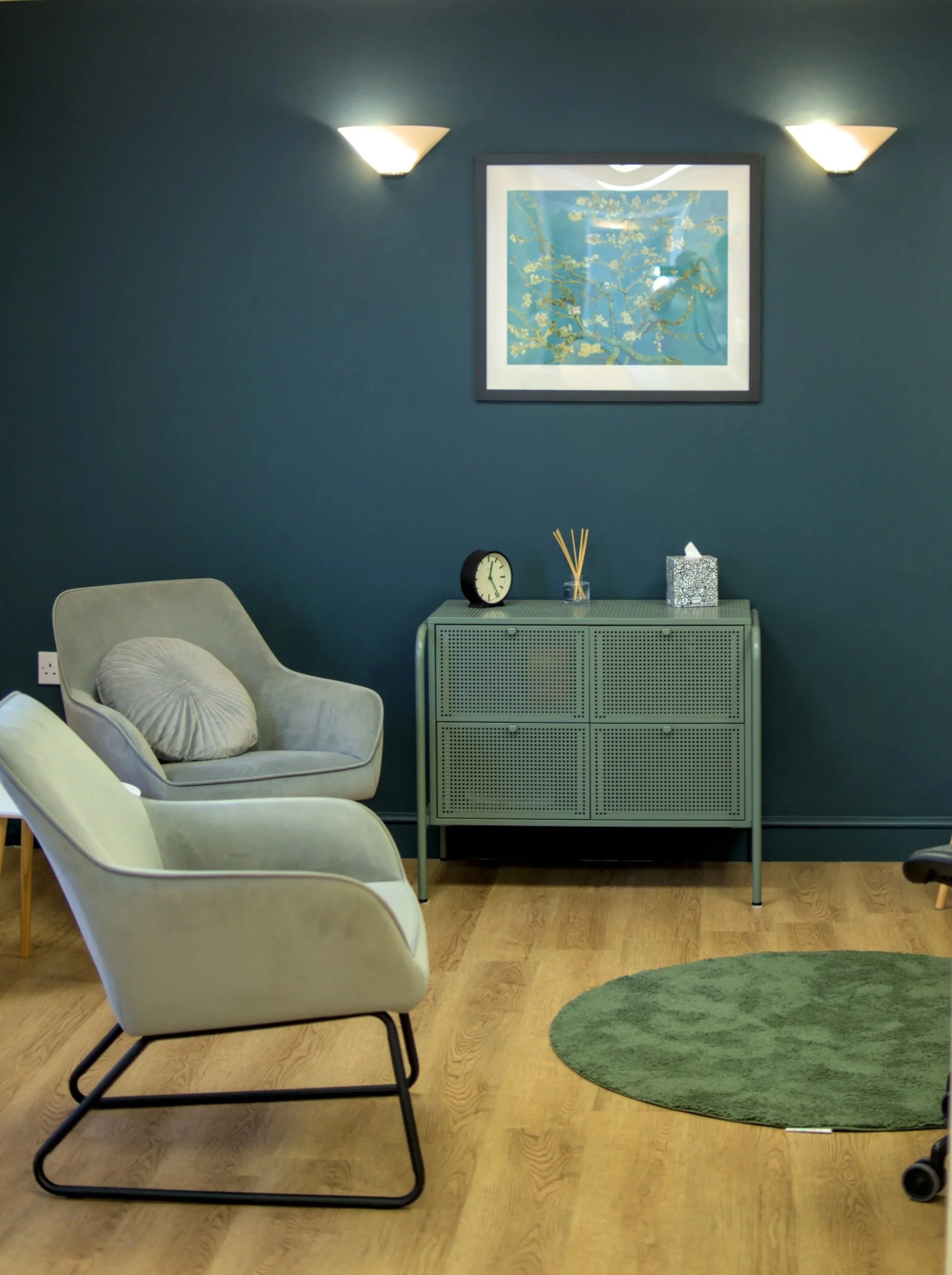 A room with white chairs, a green rug, a teal cabinet, a picture on the wall, a clock, reed diffuser, and tissue box on the cabinet.