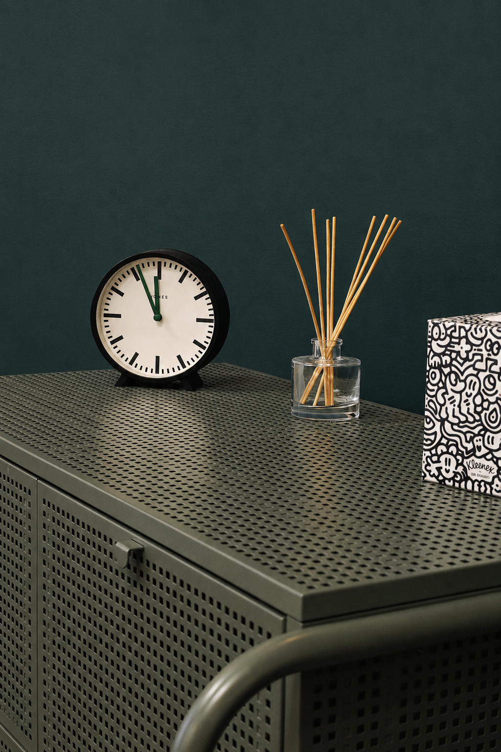 A black and white clock showing 11:55, a glass vase with bamboo sticks, and a patterned tissue box on a perforated metallic cabinet against a dark green wall.