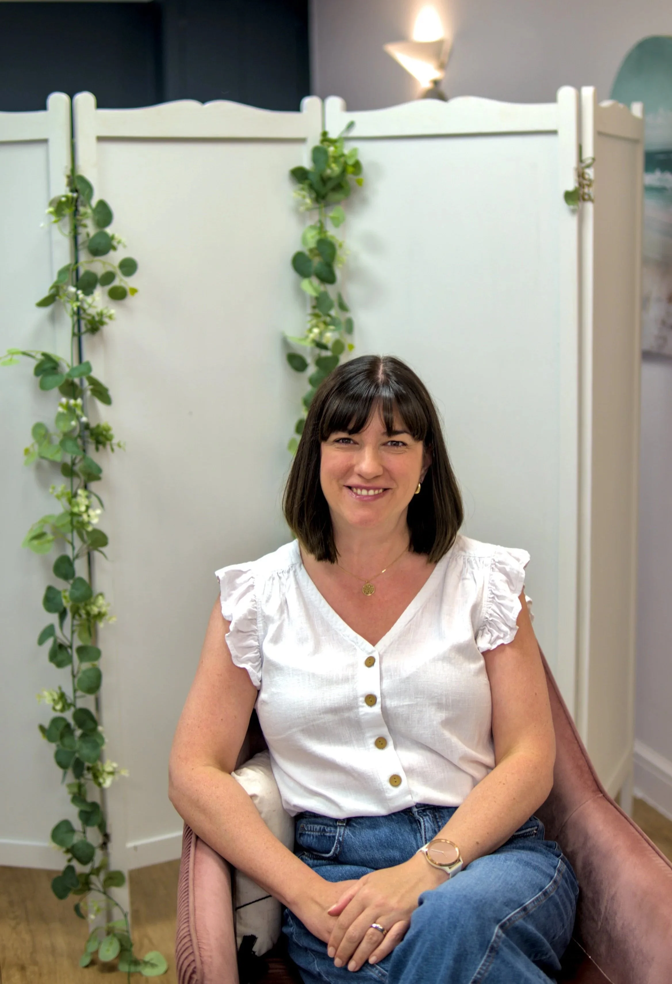 A woman with shoulder-length dark hair and a white blouse sitting on a pink armchair in front of a white room divider decorated with green ivy plants. She is smiling and wearing a gold necklace and watch.