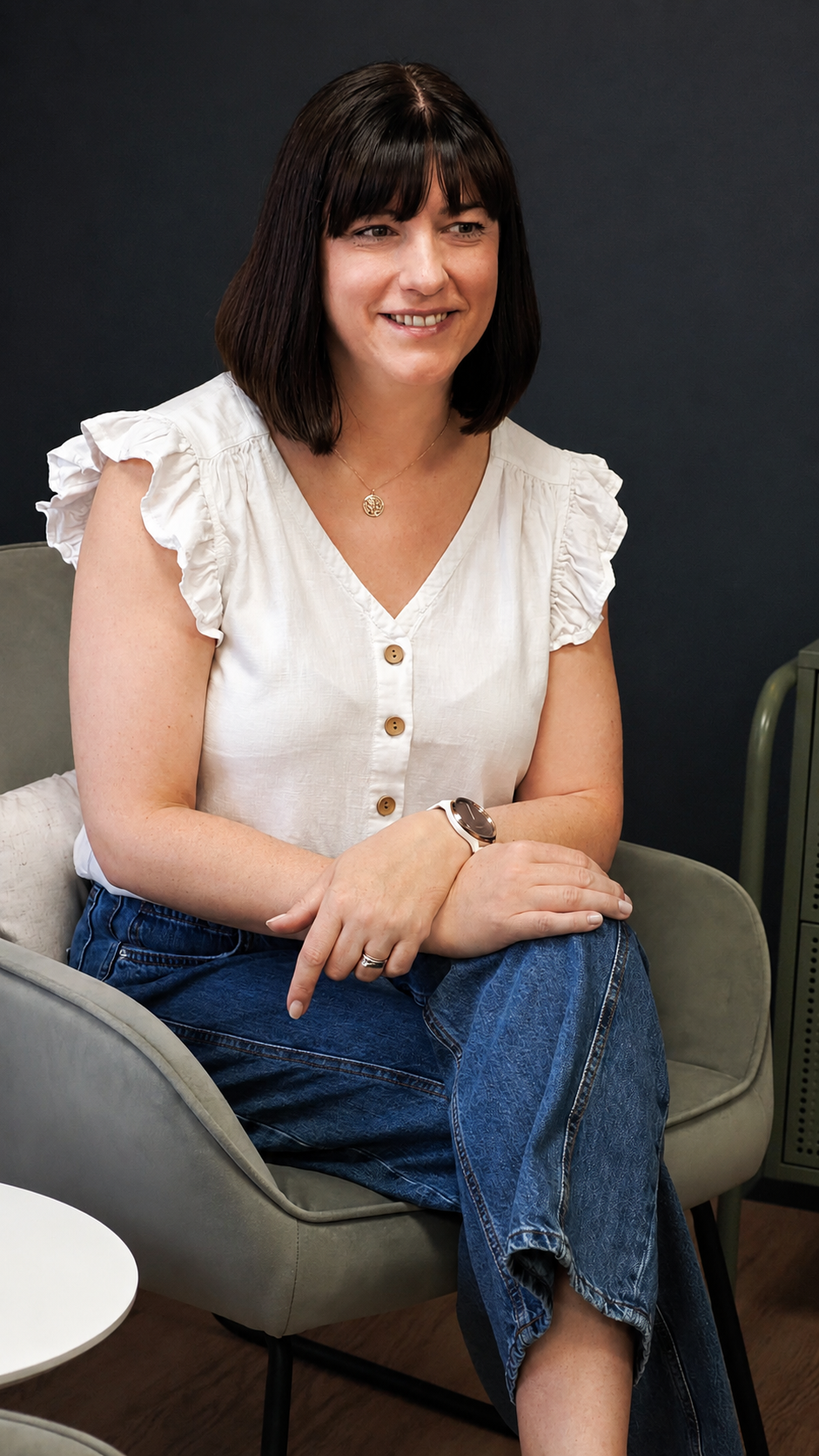A woman with short dark hair, wearing a white short-sleeved top with ruffled sleeves, sitting on a modern gray chair with her arms crossed. She is smiling and looking to her left, wearing a watch, ring, and a necklace, with a dark background behind her.
