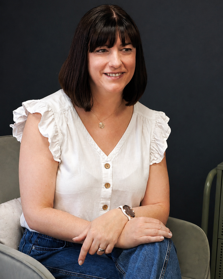 A woman with shoulder-length dark brown hair, wearing a white ruffled-sleeve blouse with buttons, blue jeans, a wristwatch, and a necklace, sitting on a gray chair against a dark background, smiling slightly.