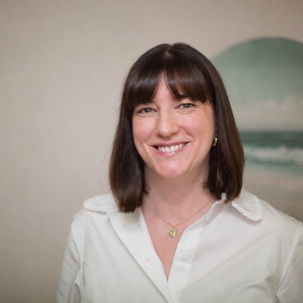 A woman with shoulder-length brown hair smiles in front of a wall with ocean artwork.