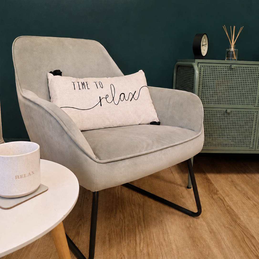 A cozy living room corner with a light gray armchair holding a pillow that reads 'Time to relax'. Next to the chair is a small white side table with a white mug labeled 'Relax'. In the background, a green cabinet with a black clock and a vase with reed diffusers on top, all against a dark green wall.