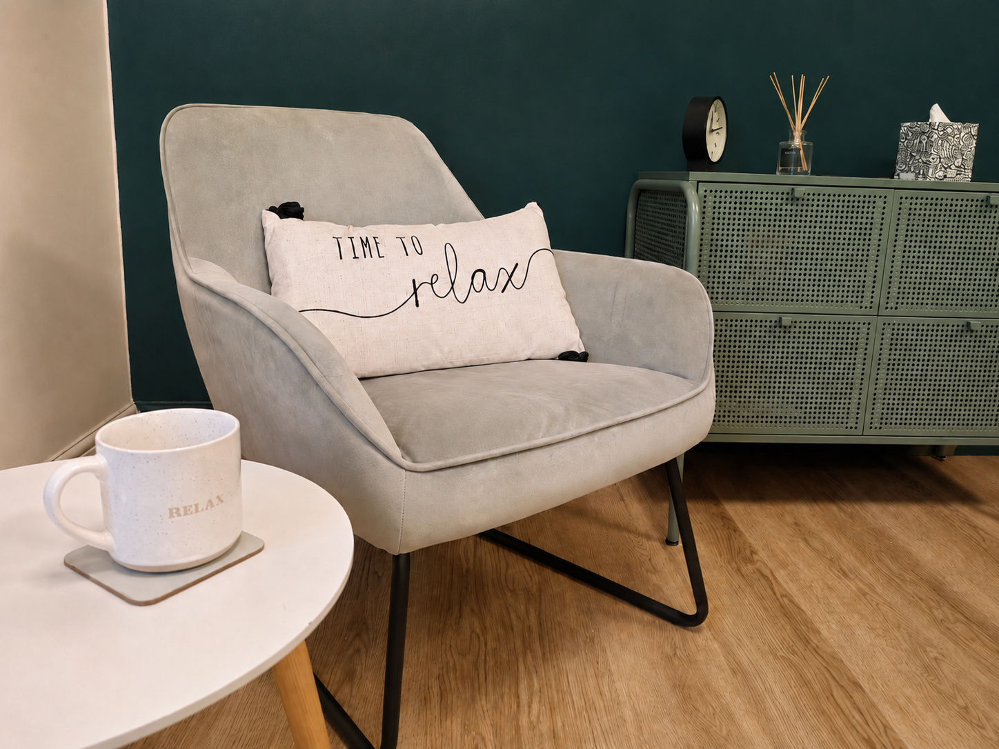 Living room corner with a beige couch, decorative pillow reading "Time to relax," a white coffee mug on a small table, and a green sideboard with a clock, diffuser, and tissue box.