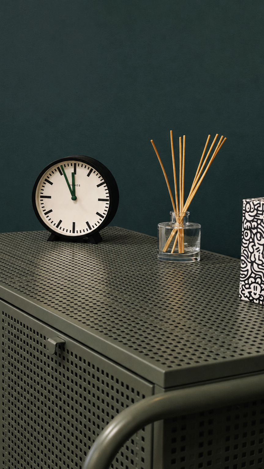 A black and white clock showing 11:55, a glass container with wooden sticks, and a patterned box on a perforated metallic surface against a dark teal wall.