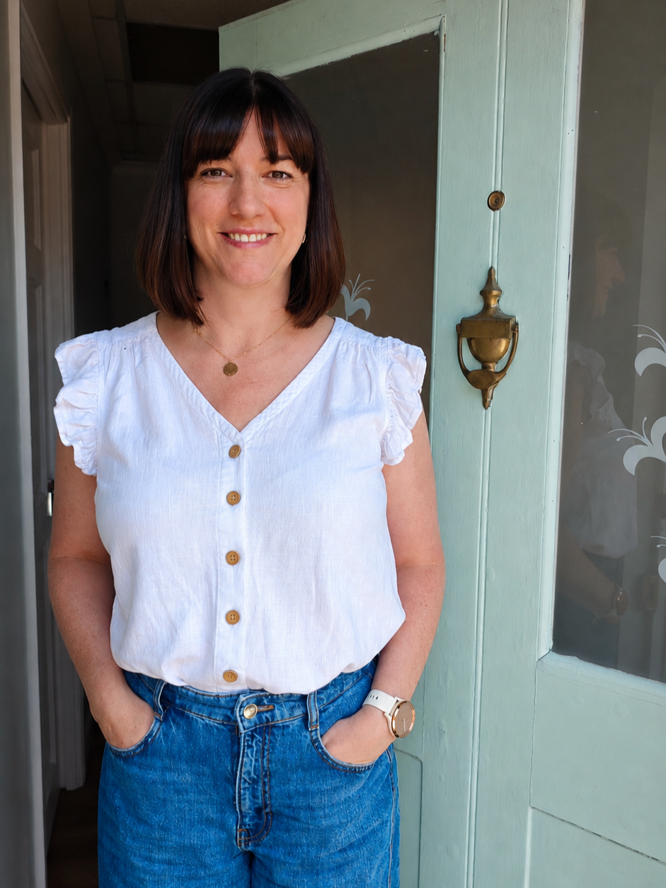 A woman with shoulder-length dark brown hair, wearing a white button-up top with ruffled sleeves, blue jeans, a gold necklace, a white watch, and small earrings, standing in front of a green door with a gold handle.