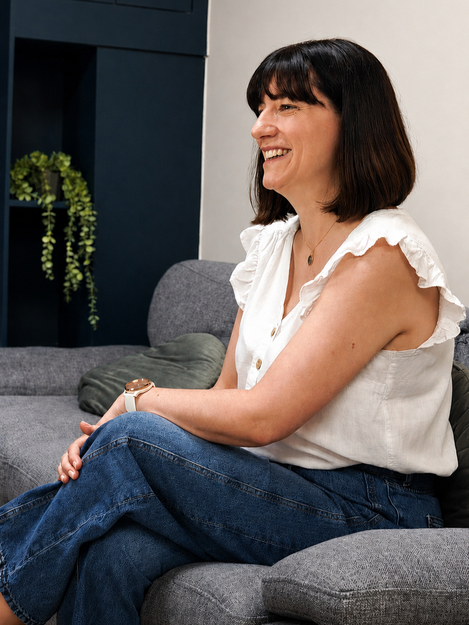 Woman with dark hair and a white sleeveless blouse, smiling while sitting on a gray sofa. There is a plant in the background.