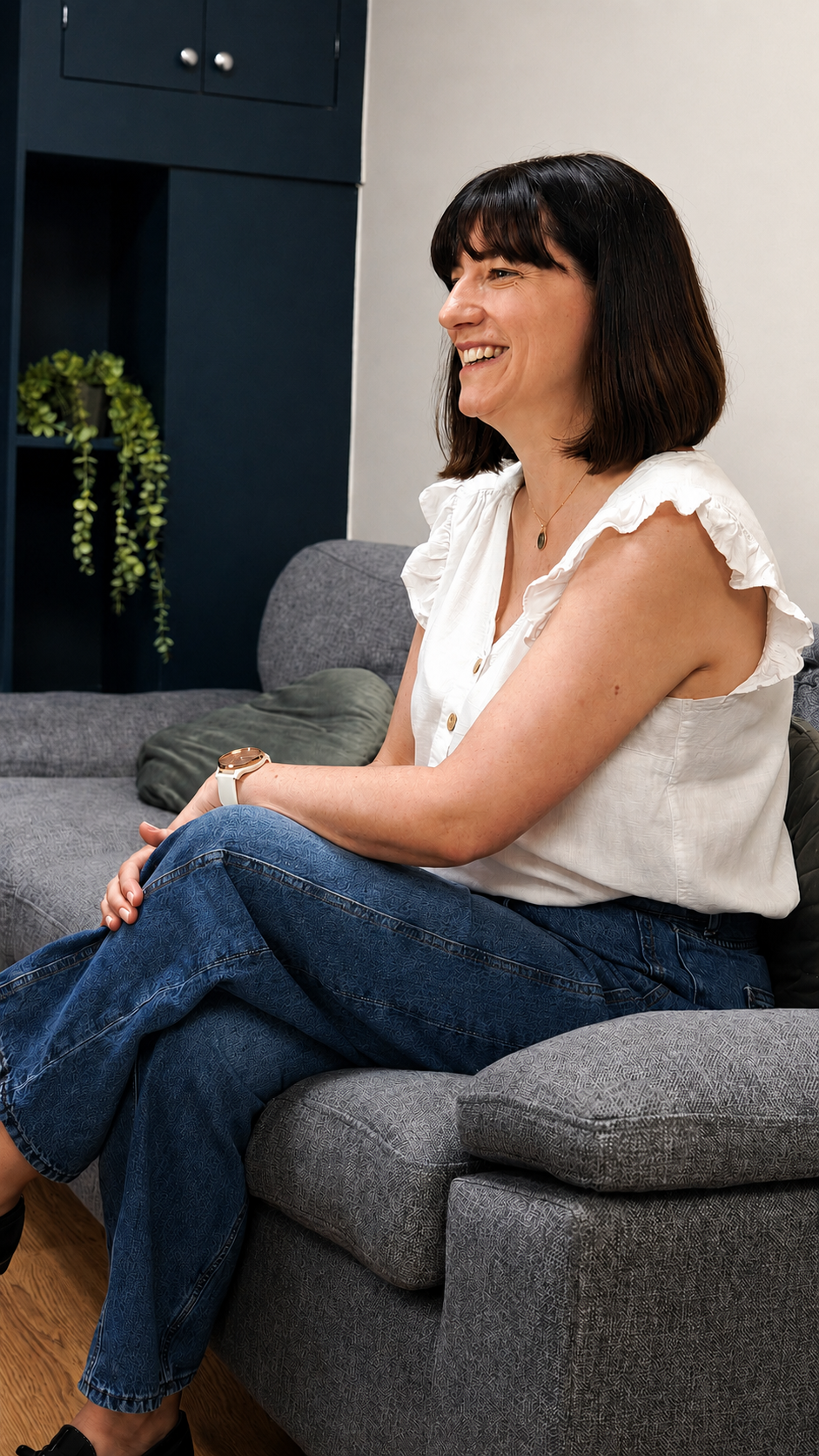Woman sitting on a gray couch, smiling, wearing a white sleeveless top and blue jeans, in a living room with a dark blue wall and a green plant in the background.