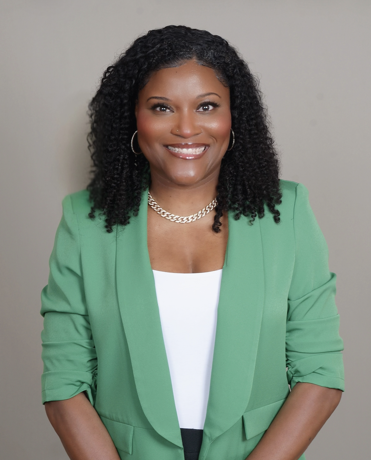 A woman with curly black hair wearing a green blazer, white top, gold necklace, and hoop earrings, smiling against a plain background.
