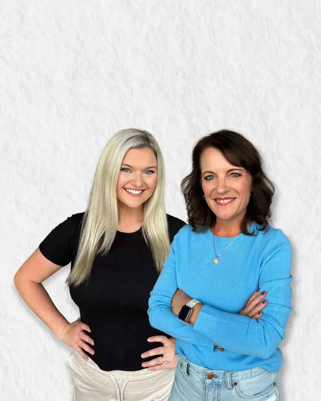 Two women standing in front of a white wall, smiling at the camera. The woman on the left has long blonde hair and is wearing a black shirt. The woman on the right has shoulder-length dark hair and is wearing a blue sweater with a smartwatch on her wrist.