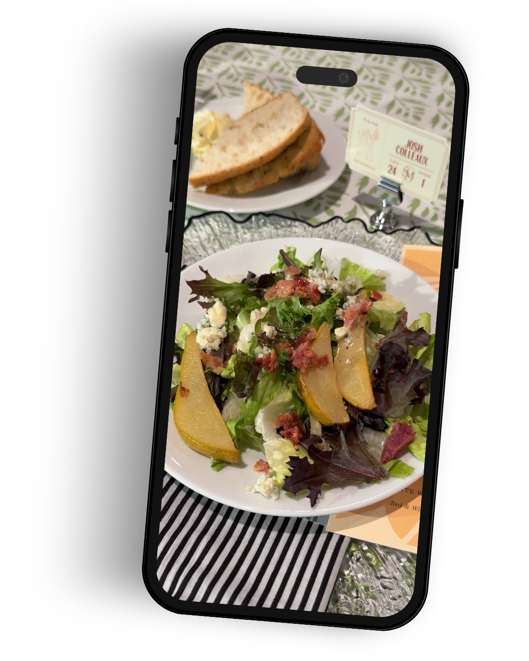A tablet displaying a photo of a salad with mixed greens, pear slices, bacon bits, blue cheese crumbles, and possibly nuts, on a table with a black and white striped cloth. In the background, there is a plate with bread and a small sign with a name and date.