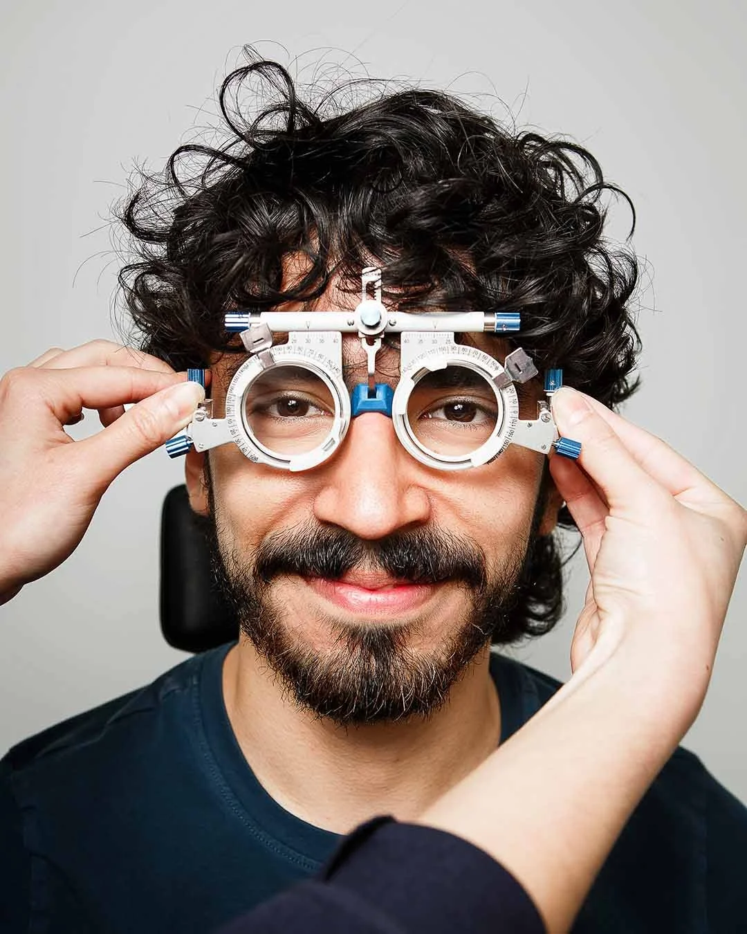 A man with curly dark hair and a beard is smiling while holding a trial frame for eye examination in front of his face.