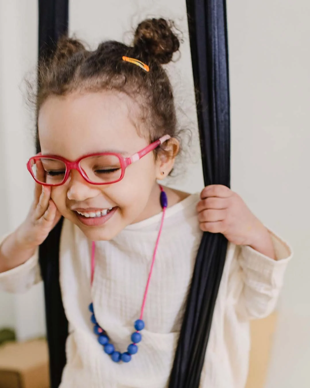 A young girl with curly hair in pigtails, wearing pink glasses, a white shirt, and a blue beaded necklace, smiling and holding onto the side of a wooden or metal object.