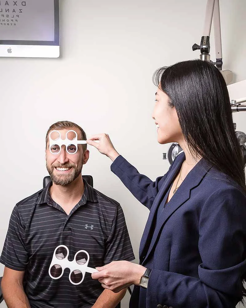 A woman with long dark hair wearing a blue blazer is holding a red rubber band and positioning a phoropter mask over a man's eyes during an eye exam in a medical office.