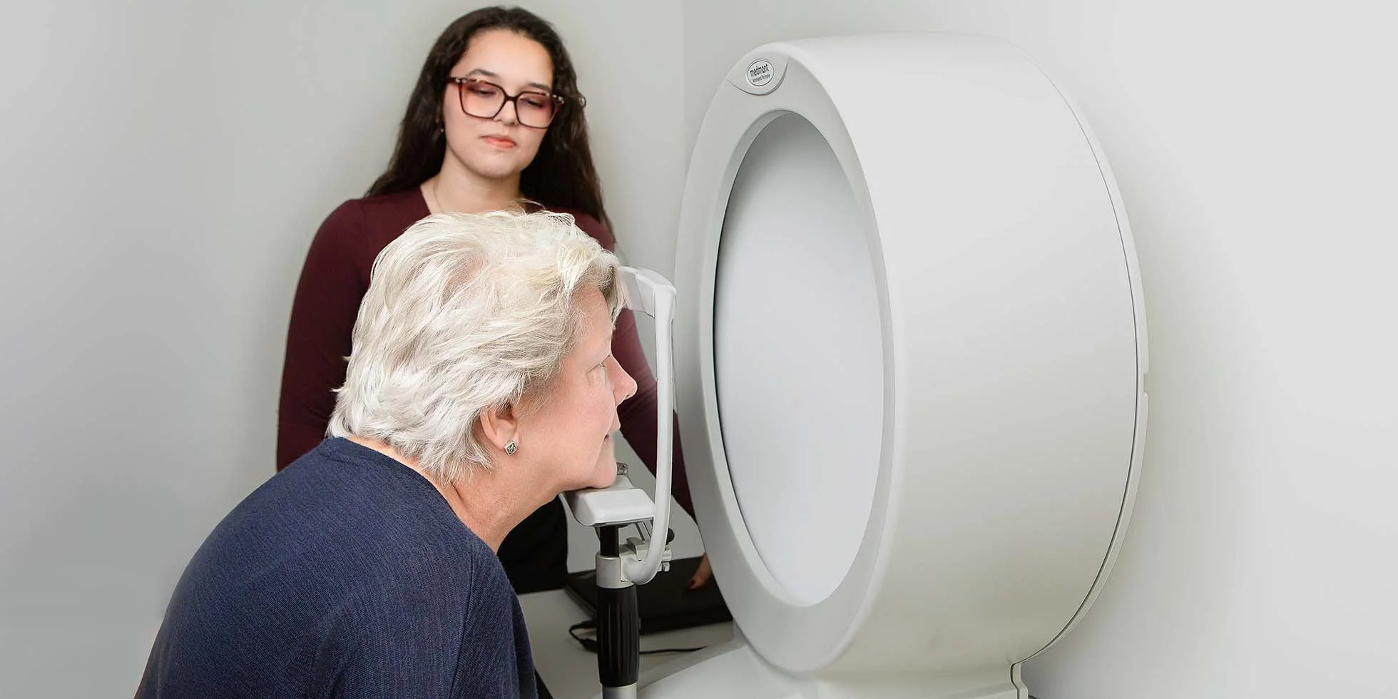 A woman is undergoing an MRI scan while another woman, likely a technician, observes. The woman to be scanned is seated with her chin on a support, looking into the enclosed MRI machine in a medical setting.