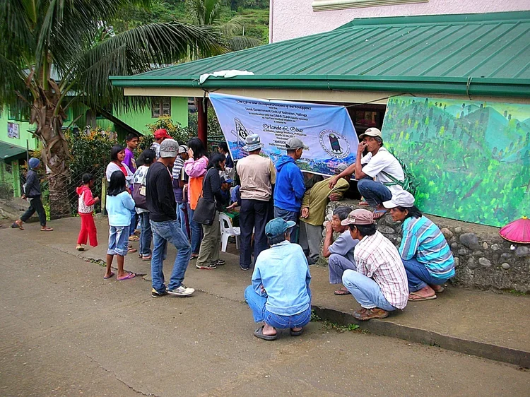 Group of people gathered outside a building with a green metal roof, some sitting and some standing, with children and adults, in a lush, hilly area, possibly attending an event or waiting in line.