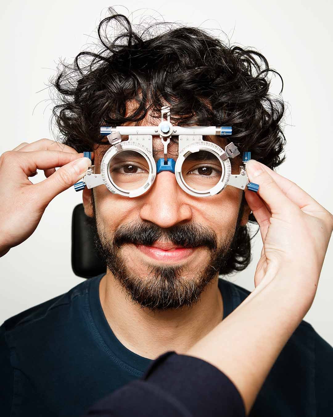 A man with curly hair and a beard wearing a blue shirt has eye examination glasses placed on his face by an optometrist.